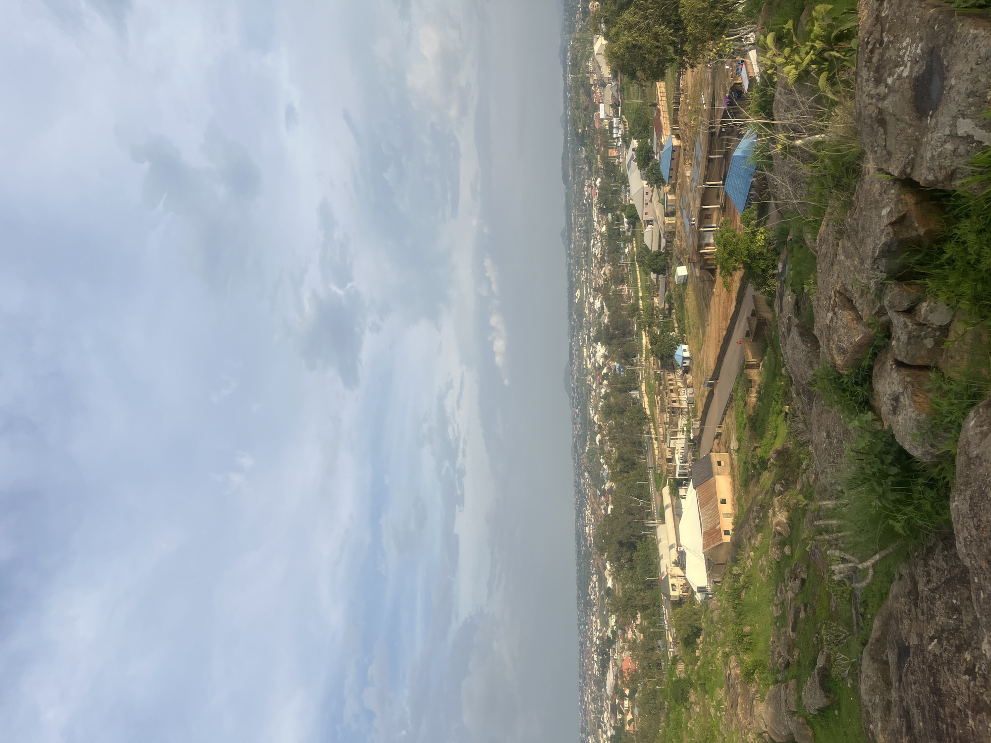An aerial, wide shot captures the sprawling urban landscape of Gura Top, Nigeria under a cloudy sky. The foreground is dominated by large, rough-hewn rocks and patches of green grass, suggesting a natural, elevated vantage point. Descending into the midground, the rocky terrain gives way to a densely populated area with numerous buildings characterized by varied roof colors, including red, white, and blue. Trees are interspersed throughout the urban area, providing green canopies. In the distance, the city stretches towards a hazy horizon, with more buildings and vegetation visible. The overall scene depicts a vibrant, developing city with both natural and man-made elements.