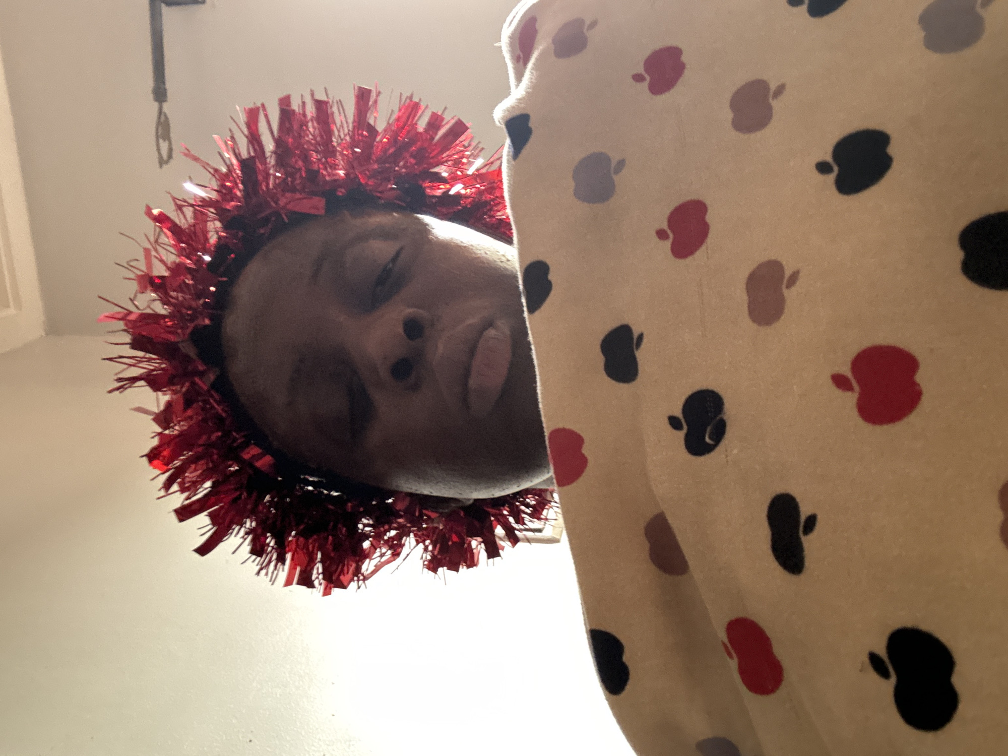 A low-angle shot depicts a person with dark skin wearing a red, fringed headband. The person's eyes are closed, and their face is angled downwards towards the camera. To the right of the person is a piece of fabric with a pattern of apples in various colors: red, black, and brown. The background is a plain white wall and a doorway in the upper left corner. The photograph does not provide any specific details linking it to Sabon Tasha, Nigeria.