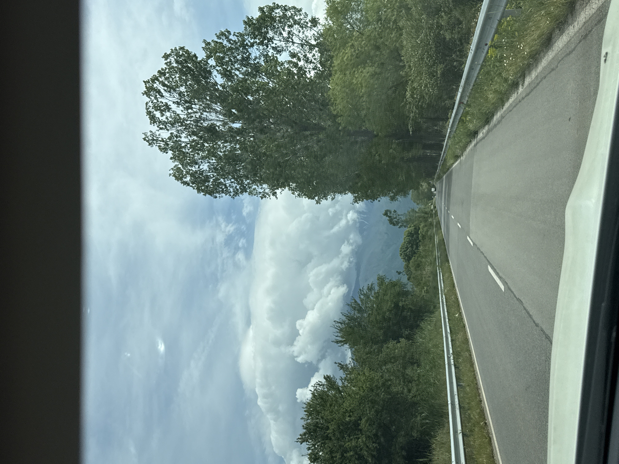 The image shows a road in Kocherinovo, Bulgaria, from the perspective of a vehicle. The road is asphalt, marked with white lines, and has a metal guardrail on the side. Trees line the road. In the background, there are mountains and a cloudy sky. The image appears to be taken through a car window, as part of the car's frame is visible.