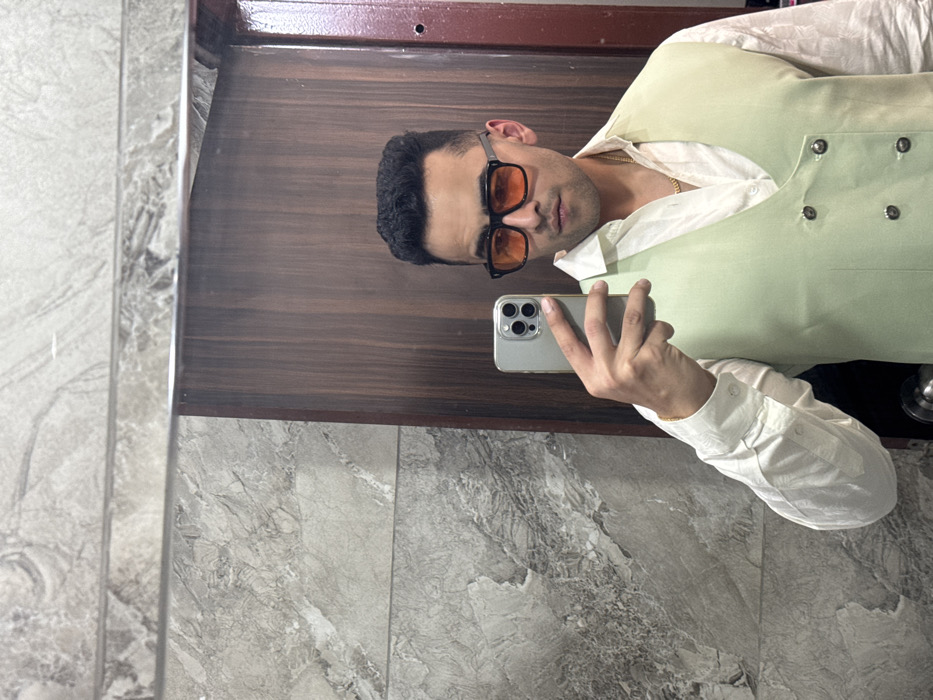 An individual is depicted taking a mirror selfie indoors in Malasar, India. The subject is a young adult male, likely of South Asian descent, with short, dark hair. He is wearing a white collared shirt, unbuttoned at the top to reveal a gold chain, and a light green double-breasted vest with dark buttons. His eyes are obscured by black-framed sunglasses featuring orange-tinted lenses. He holds a silver-colored smartphone, equipped with a clear protective case, in his right hand, angled towards the mirror to capture his reflection. The background behind the subject, as seen in the mirror, consists of light gray marble-patterned wall tiles. A horizontal reflective strip divides these tiles. To the right, a vertical section of dark brown wood paneling or a door frame is visible.