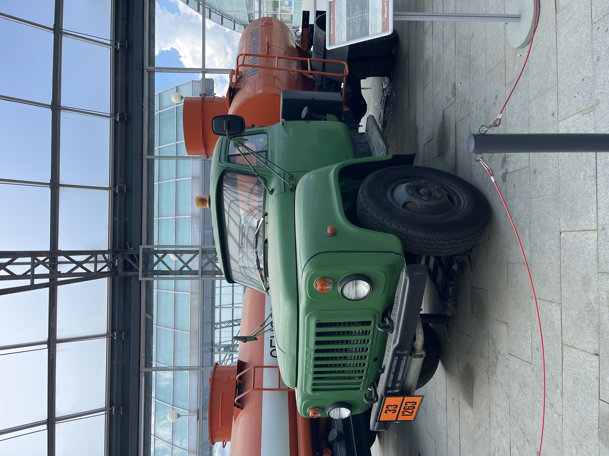 In the city of Verkhnyaya Pyshma, Russia, a green GAZ-53 fuel truck is parked indoors under a modern glass and metal structure. The truck, with its distinctive orange hazard plate displaying "33 1263", is positioned in front of a large orange fuel tanker. The tanker has Cyrillic lettering on its side. The scene appears to be a museum exhibit, with a stanchion and red rope cordoning off the area. The floor is paved with light-colored square tiles, and reflections of the cloudy sky are visible on the glass ceiling.