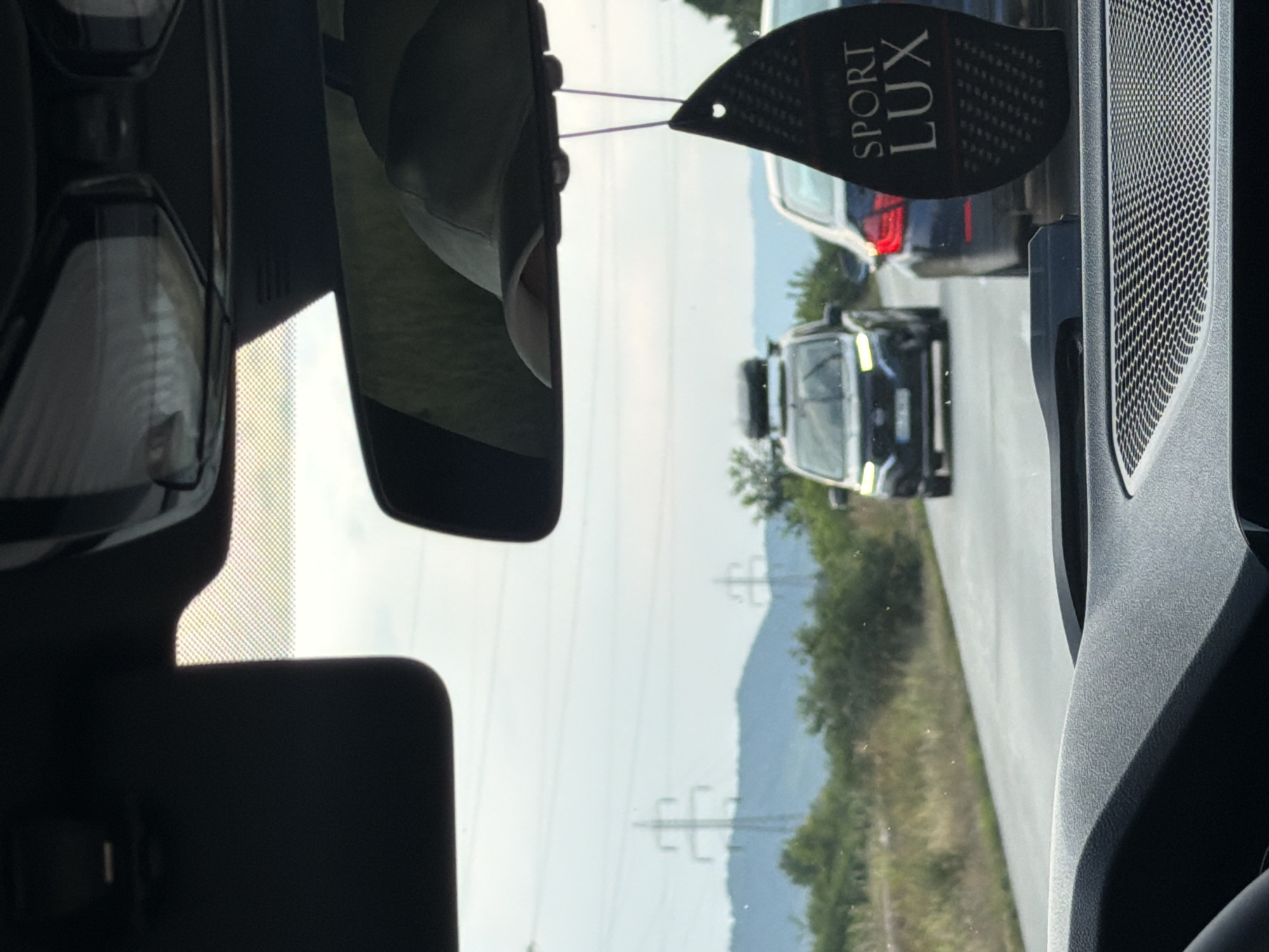 The image, taken from the interior of a vehicle, depicts a traffic scene on a paved road. In the foreground, the dashboard and part of the windshield are visible, along with a black air freshener hanging from the rearview mirror. The air freshener is oval-shaped with a textured surface and the words "SPORT LUX" printed on it.
Further down the road, a dark-colored van with a roof rack is visible, followed by a lighter-colored car. The van's headlights are on. Both vehicles are in motion, suggesting the image was taken while driving.
The surrounding environment consists of a roadside with dry grass and vegetation. In the background, a line of trees and hills can be seen, with power lines and transmission towers indicating infrastructure in the area. The sky is overcast, suggesting it may be a cloudy day.
The location is identified as Onești, Romania, based on contextual information provided. The scene is typical of a road in a semi-rural or suburban area, potentially on the outskirts of the city, given the presence of natural landscape alongside road infrastructure. No people are clearly visible within the vehicles or outside of them.