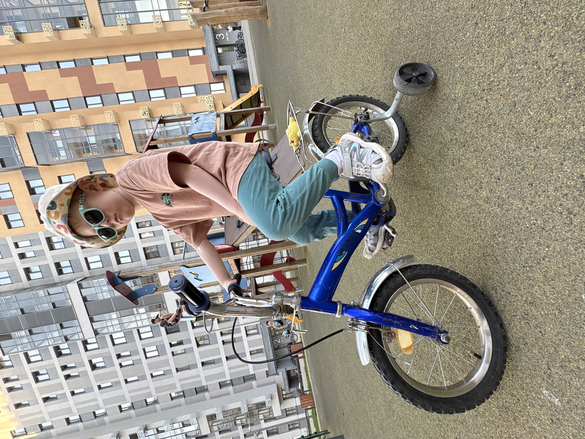 A child in a pink t-shirt, teal pants, a patterned hat, and sunglasses is riding a blue bicycle with training wheels. The child is on a playground in Izhevsk, Russia, with modern apartment buildings in the background. A seesaw and other playground equipment are visible. The ground is a textured, greenish-brown surface.
