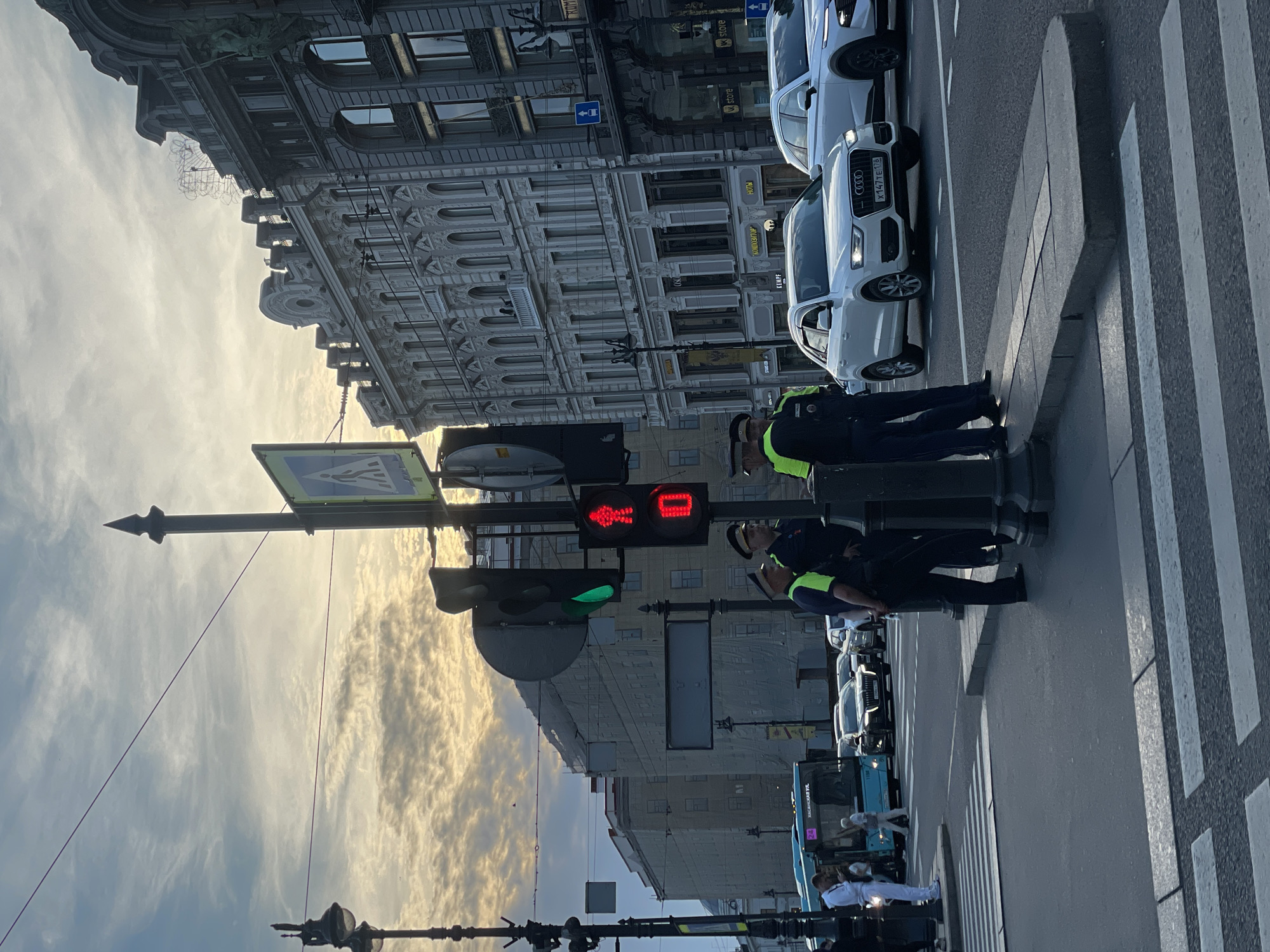 The image captures a vibrant street scene in Saint Petersburg, Russia, during what appears to be the late afternoon or early evening, characterized by warm, soft sunlight illuminating dramatic clouds in the sky.
In the foreground, two uniformed traffic policemen stand on a paved sidewalk, positioned near a crosswalk. The pedestrian traffic light above them displays a red "stop hand" symbol and a red "walking man" countdown, indicating that pedestrians should not cross. Conversely, the adjacent vehicle traffic light is green, allowing traffic to proceed.
A line of vehicles is visible on the asphalt road, including several white cars, one of which is an Audi SUV clearly displaying a Russian license plate "X147TE 178," with the "178" region code confirming Saint Petersburg. Further down the street, a light blue bus and other pedestrians can be seen.
The street is flanked by imposing, multi-story buildings with intricate, ornate architectural details, featuring numerous windows and decorative facades typical of European city centers. Several signs with Cyrillic text are visible on these buildings, including "РУНЫ" (Runes), "K_STORE", "N_D", "КОНДИТЕРСКАЯ" (Confectionery), and "УНД". A pedestrian crossing sign is also visible overhead. The composition offers a glimpse into the bustling urban environment of Saint Petersburg, with its distinctive architecture and everyday street activity.