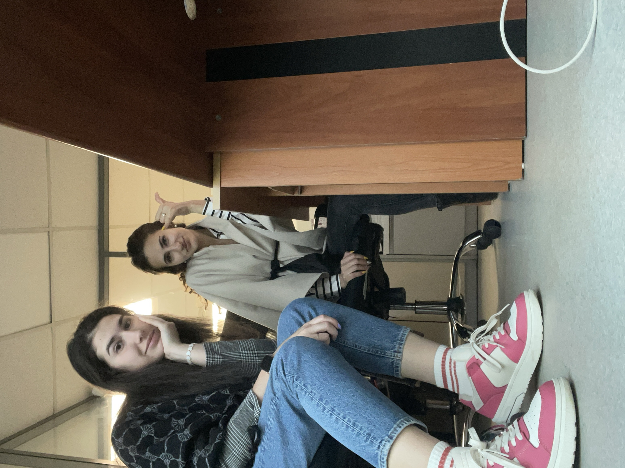 Two young women are sitting on rolling chairs under a wooden desk. One woman is closer to the foreground, wearing blue jeans, red and white sneakers, a black and white patterned top, and a beaded bracelet. She is resting her head on her hand. The other woman is behind the desk, wearing a beige jacket and jeans. She is giving a thumbs-up. The location appears to be indoors, possibly a study room. The city and country are provided as location information (Sofia, Bulgaria), but there are no discernable features within the image to confirm this.