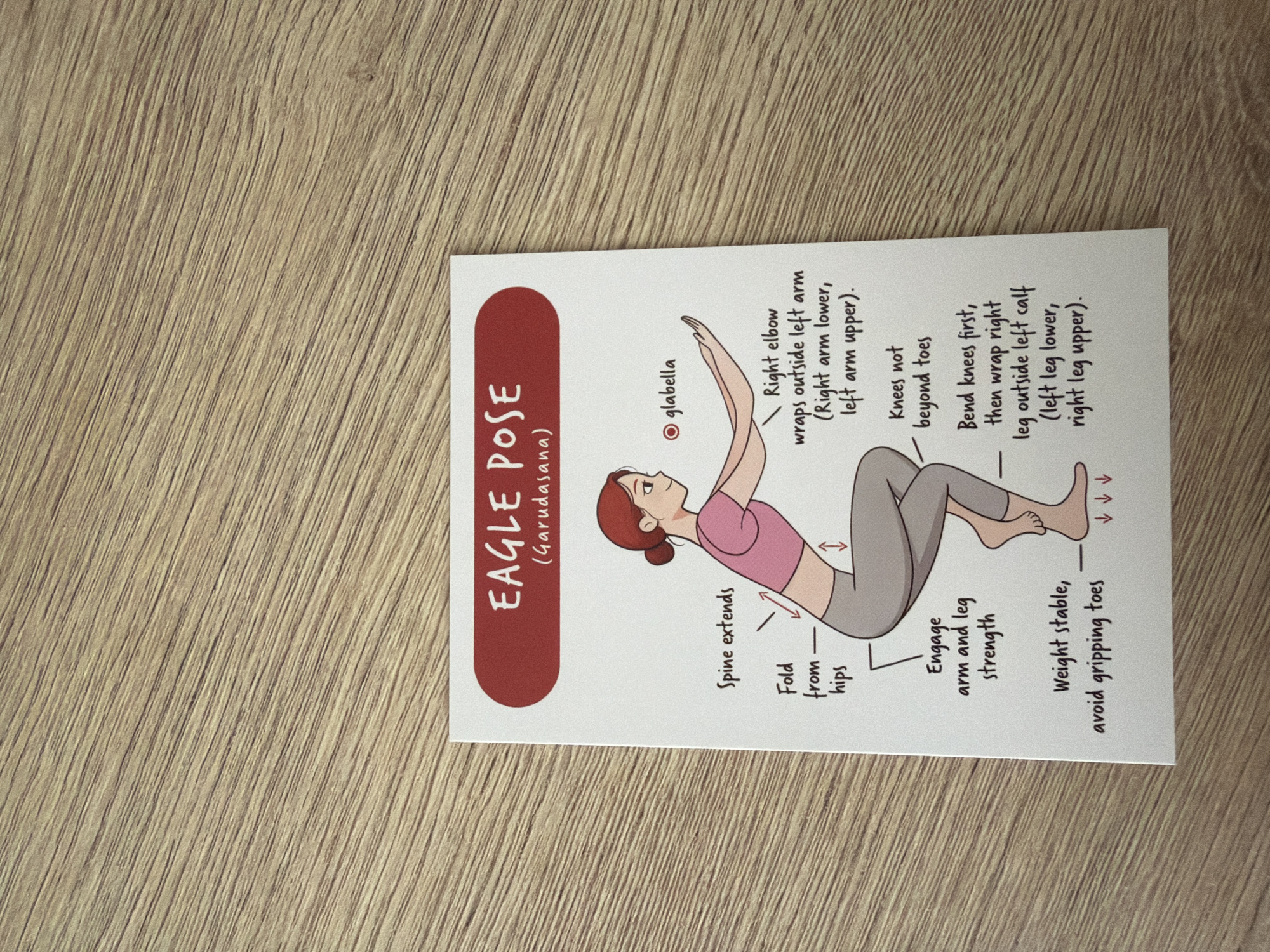 The image shows a yoga pose card titled "EAGLE POSE (Garudasana)". The card features an illustration of a woman performing the pose, with labels pointing to various parts of her body and instructions for the pose. The card is placed on a wooden surface with a visible wood grain pattern. There is no information in the image that can be used to determine its location in Pazardzhik, Bulgaria.