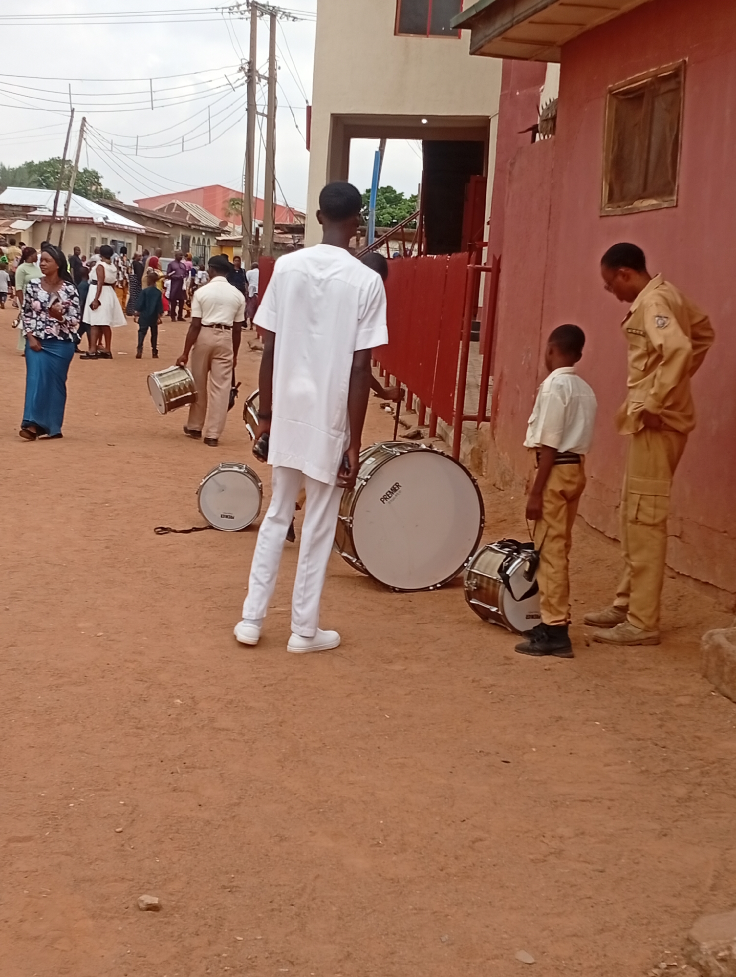 The image shows a group of people and percussion instruments in what appears to be a street in Narayi, Nigeria. The ground is dirt. In the foreground are three males and various snare and bass drums. One male wears a white shirt and white pants, while another male appears to be a young boy in a short-sleeved white shirt and khaki pants. A third male stands near the young boy; he is wearing a khaki shirt and pants. Several other people are walking in the background of the image. Behind them are other buildings. Overhead are power lines.