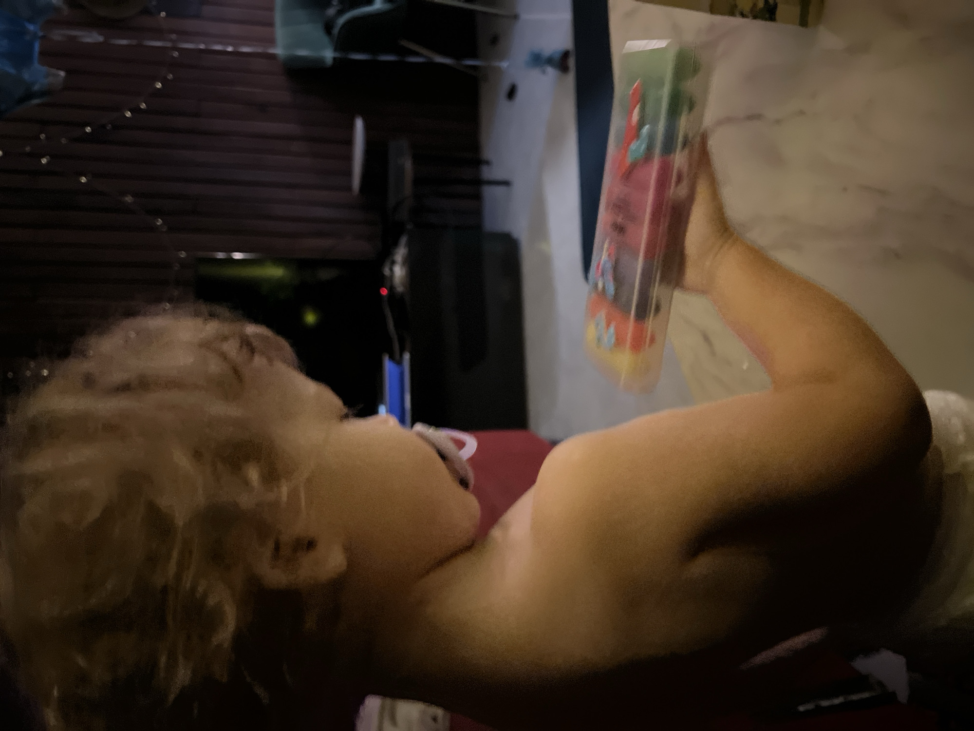 A toddler with blonde, curly hair is shown from the side, wearing a pacifier. They are holding a clear plastic case filled with colorful objects. The background is dark and out of focus, with some vertical wooden planks and string lights visible. There is a table and chair in the background, and a hint of a television screen. The scene appears to be indoors, possibly in a home setting.