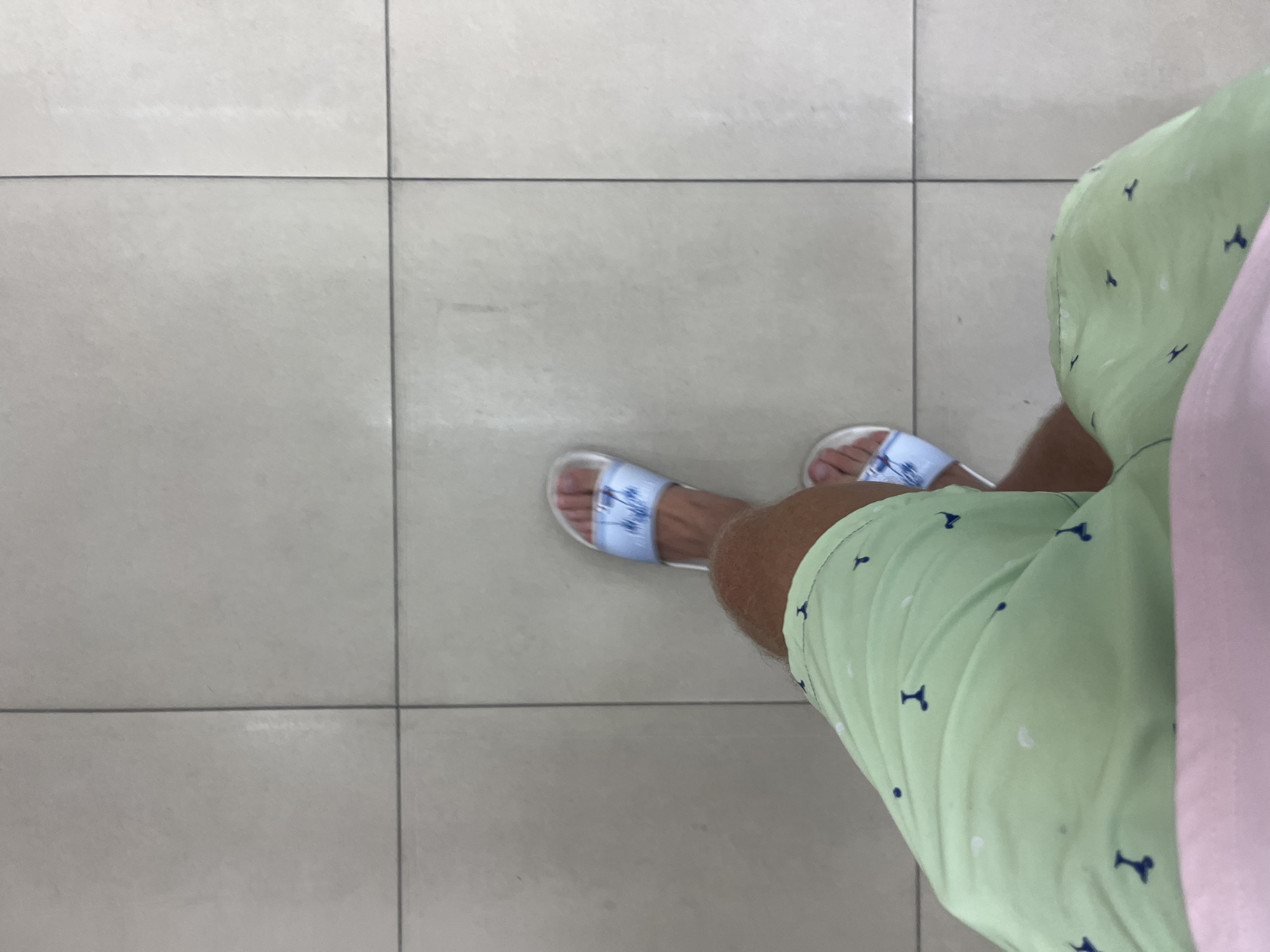 A person's legs are shown from a high-angle perspective, standing on a tiled floor. The individual is wearing light green shorts with a pattern of small, dark blue shapes that resemble anchors or stylized letters, and white polka dots. On their feet are light blue slides with a white pattern of what appears to be birds or a similar motif. The background is a floor composed of large, light-colored square tiles with dark grout lines. The scene appears to be indoors, possibly in a commercial establishment or residence, given the uniform tiling. The image does not provide any specific visual cues to determine the exact location within Rawai, Thailand.