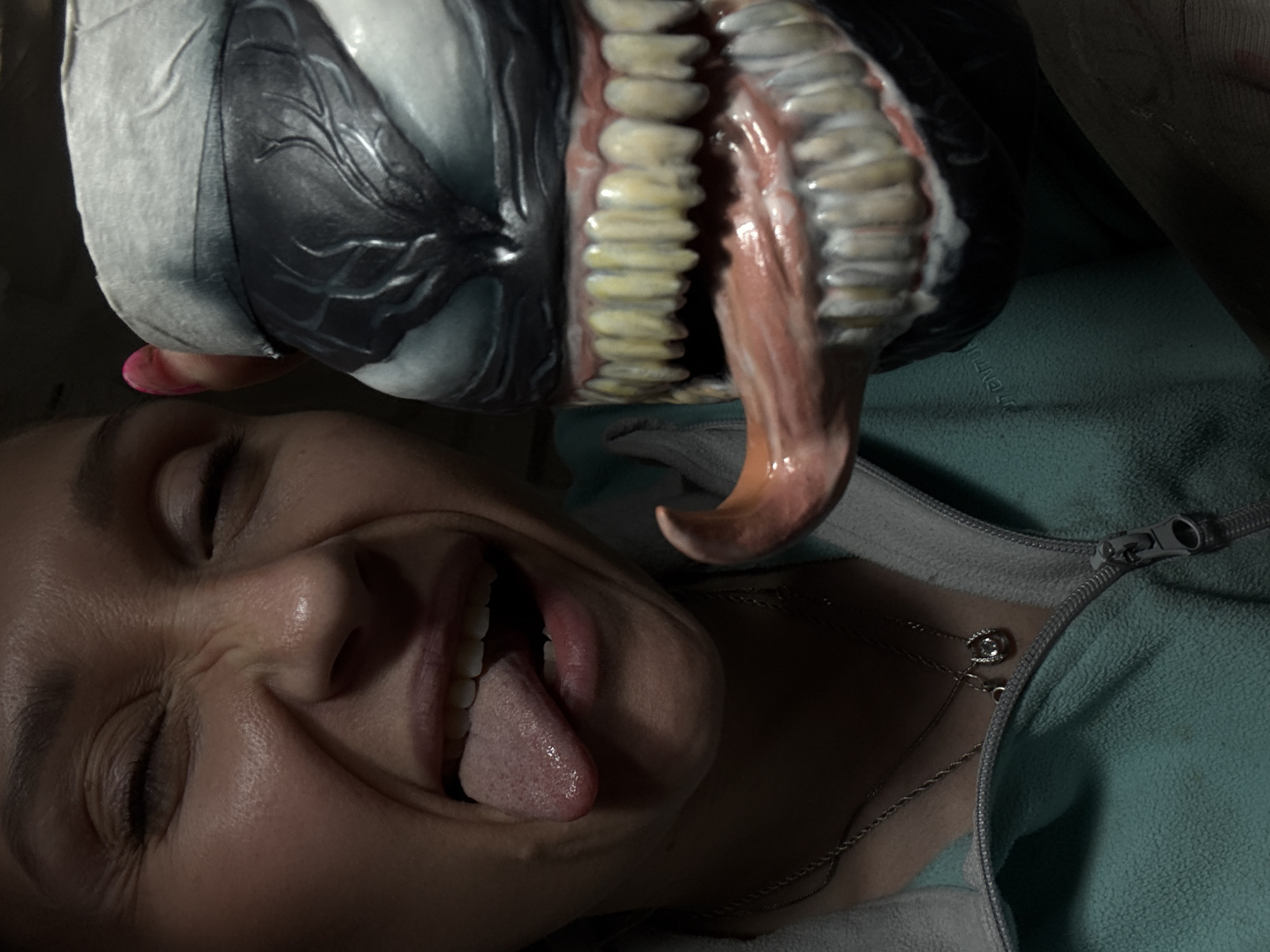 A woman is lying on her back, wearing a teal zippered jacket and a silver necklace with a circular pendant. She is sticking her tongue out and smiling with her eyes closed. Above her is a Venom mask, held in place by a pink fingernail, with its mouth open and a long, pink tongue protruding. The image does not provide enough information to determine the location in the city of Chaikovsky, Russia.
