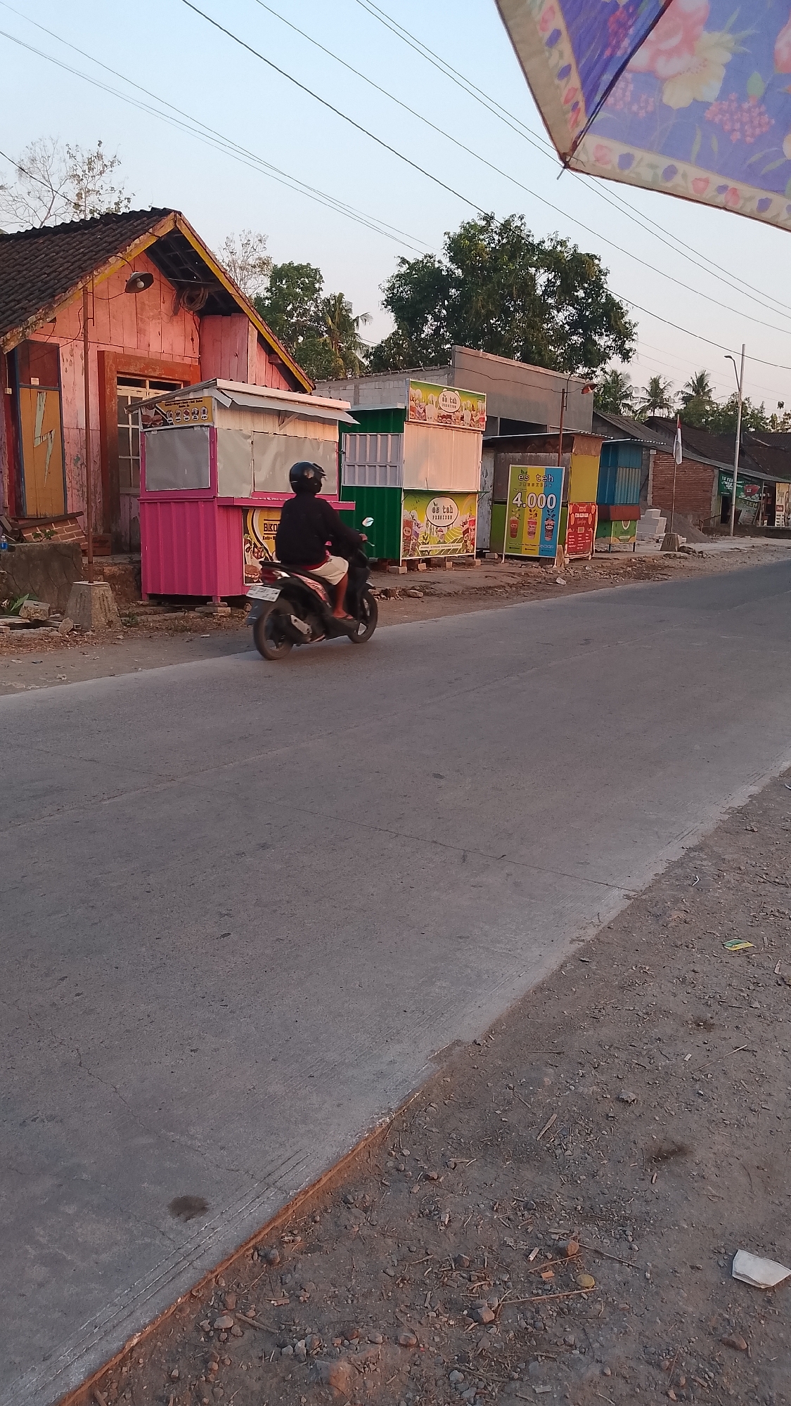 A person wearing a black helmet and a black long-sleeved shirt is riding a black and white motorcycle on a paved road. They are moving from left to right in the frame. In the background, there are several small colorful structures that appear to be food stalls. One stall is bright pink, another is green, and there are several white stalls with signage. Behind the stalls, there is a pink wooden building with a dark roof. Trees are visible behind the buildings and stalls. A portion of a colorful umbrella is visible in the top right corner of the image. The sky is a pale blue, suggesting it is either early morning or late afternoon. The scene appears to be in a rural or semi-urban area along a road.