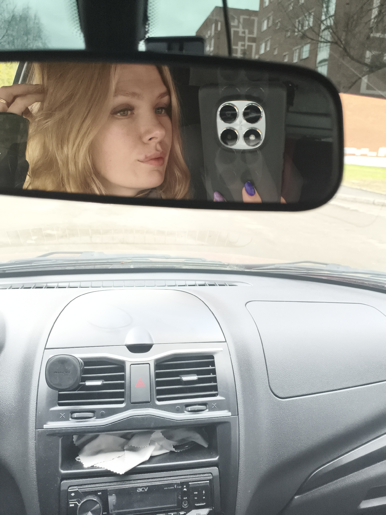 The image provides an interior view of a car, seen from the perspective of a driver or front passenger. The main focus is the car's rearview mirror, which reflects a young blonde woman taking a selfie.
**Subjects:**
A young woman with wavy blonde hair and long eyelashes is the primary subject, seen in reflection. She is wearing a ring on her right hand, which is visible near her hair, and her fingernails are painted a vibrant purple. She is holding a black smartphone with a distinctive quad-camera module featuring four silver-ringed lenses arranged in a square. The car's dashboard is prominent below the mirror, made of grey and black plastic. It features horizontal air vents, a red triangular hazard light button, and a black circular phone mount attached to one of the vents. Below the vents is a car stereo unit, and a small cubbyhole underneath holds a white, folded cloth.
**Scene and Setting:**
The scene is set inside a car, looking forward through the windshield. The background visible through the windshield and reflected in the mirror includes a large, multi-story building with numerous windows, suggesting an urban or suburban environment. Trees with bare branches are also visible, along with glimpses of a road and grassy areas. The overall setting is a car interior with a view of an outdoor urban landscape.
**Activity:**
The woman is engaged in taking a selfie, using her smartphone and the rearview mirror as a reflective surface. She appears to be posing, with her hand near her hair.
**Notable Details:**
The time of day is daytime, with diffused lighting suggesting an overcast or cloudy sky. The woman has a neutral expression with a slight pout. The car's dashboard appears functional. Visible text includes "BOROFONE" on the phone mount and "acv" on the car stereo, along with "AVG-1400R," "USB," "SD," "AUX," "FLAC," "IR," "SET," and "BND" on the stereo unit. The background building, visible in the mirror, is a typical residential or administrative structure found in urban settings like Izhevsk, Russia.