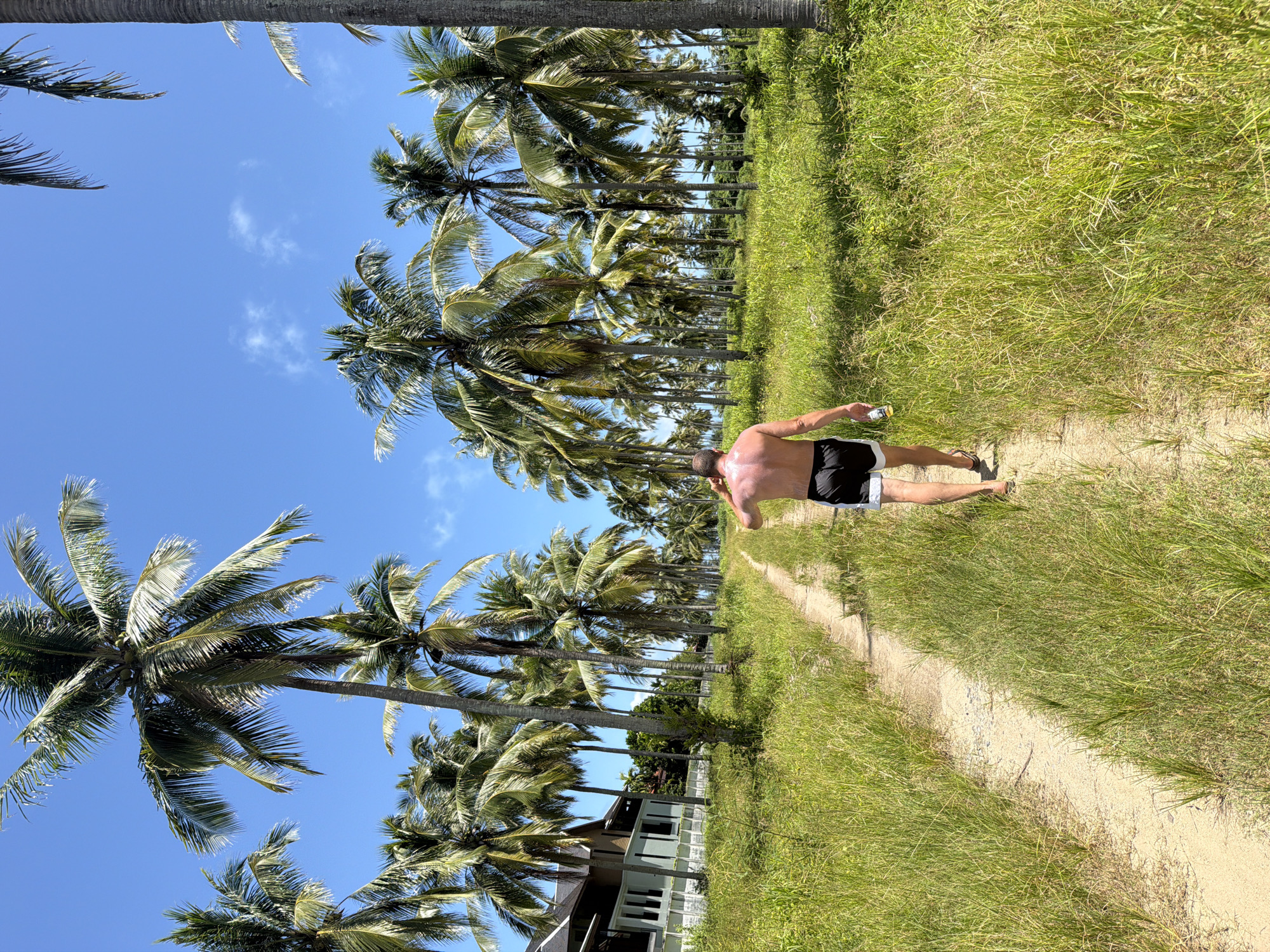 In Bang Muang, Thailand, a shirtless man walks away from the camera down a dirt path lined with tall grass and numerous palm trees. He is wearing black swim trunks with white trim and flip-flops, and he is holding a can in his right hand. The sky is a clear blue with a few scattered clouds. To the left of the path, a light-colored building with dark windows and a dark roof is visible among the trees. The scene appears to be a rural or tropical setting.