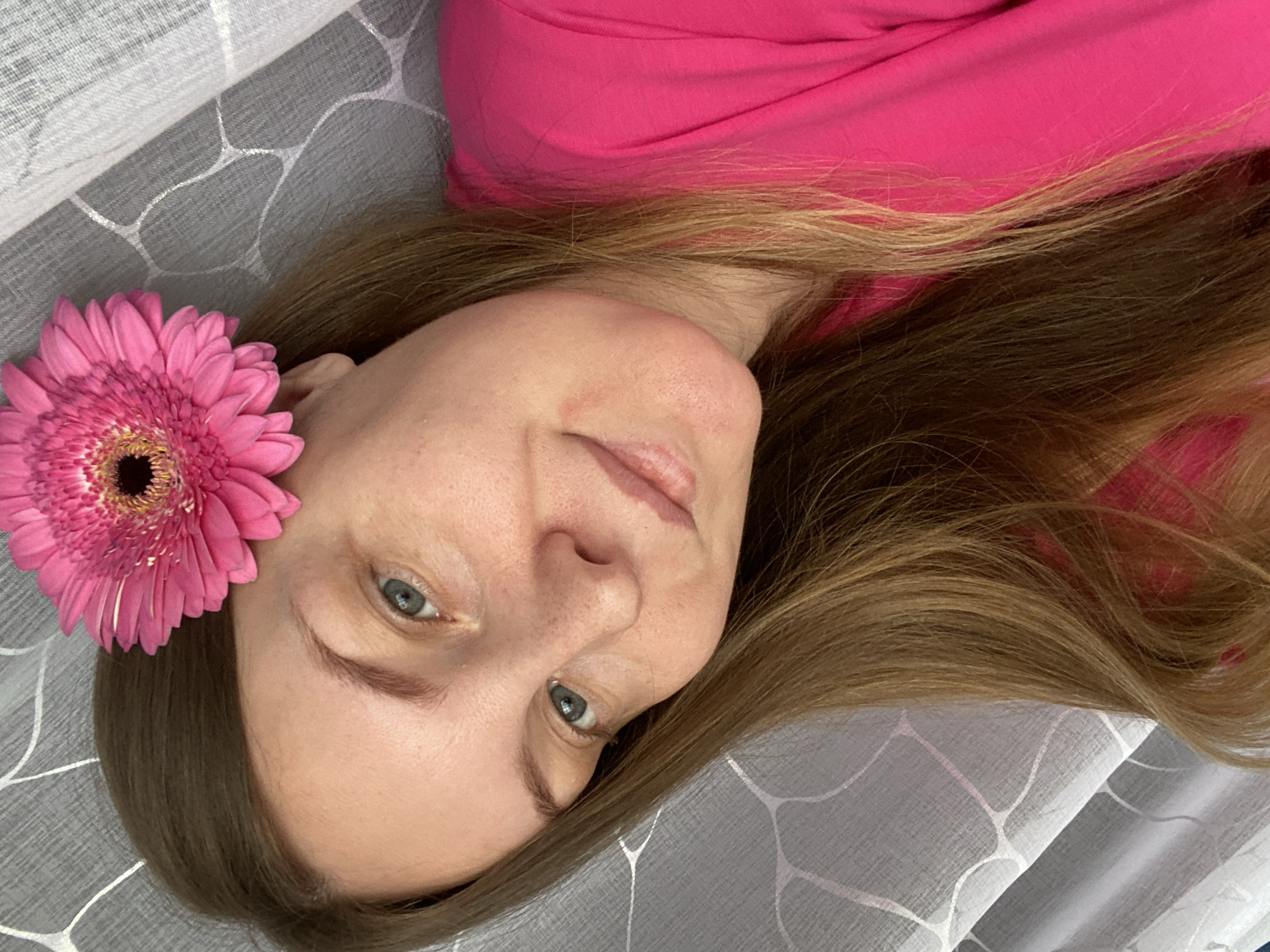 A woman lies on a gray patterned fabric. She wears a bright pink top. A pink gerbera daisy is near her ear. Her hair is brown with blonde highlights.