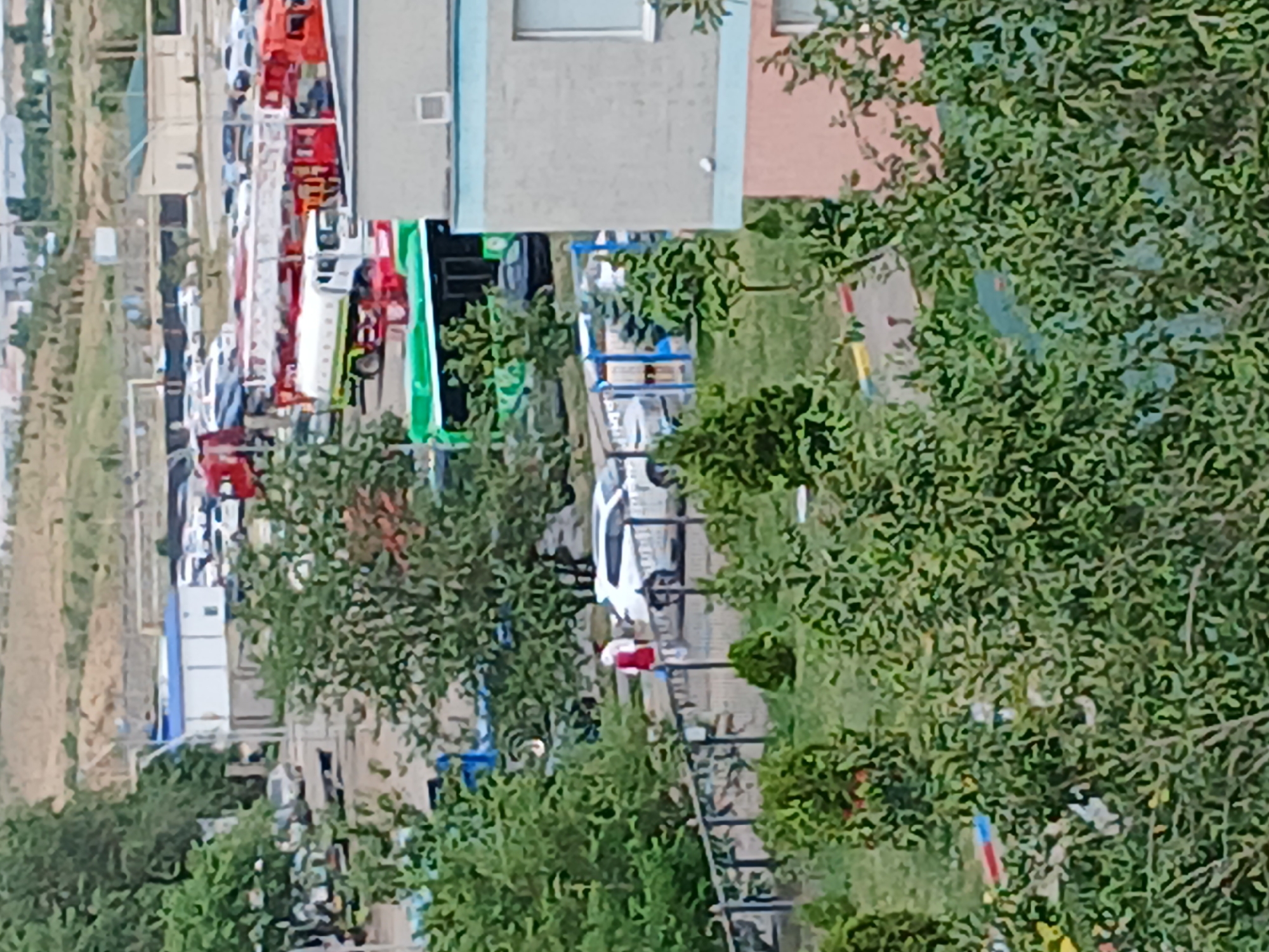This is a telephoto, landscape-oriented photograph taken from a high vantage point, overlooking a complex urban scene in Almaty, Kazakhstan. The foreground is dominated by dense green foliage, suggesting the presence of trees or bushes. In the middle ground, a variety of vehicles are visible, including several fire trucks, a white bus with green accents, a white sedan, and what appears to be a water truck. Two individuals are seen near the white sedan, one wearing a red shirt and white pants and the other in a white shirt. A black vehicle is also parked near the bus.
Further in the background, there are low-rise buildings, including a white structure with a blue roof, and beyond that, a more distant landscape with undeveloped hills or plains. The overall impression is that of a functional area, possibly a depot or an industrial zone, with a mixture of parked vehicles and some limited human activity. The lighting suggests a daytime scene, with no discernible shadows indicating a specific time of day. The image is characterized by a moderate level of detail due to the telephoto lens and distance.