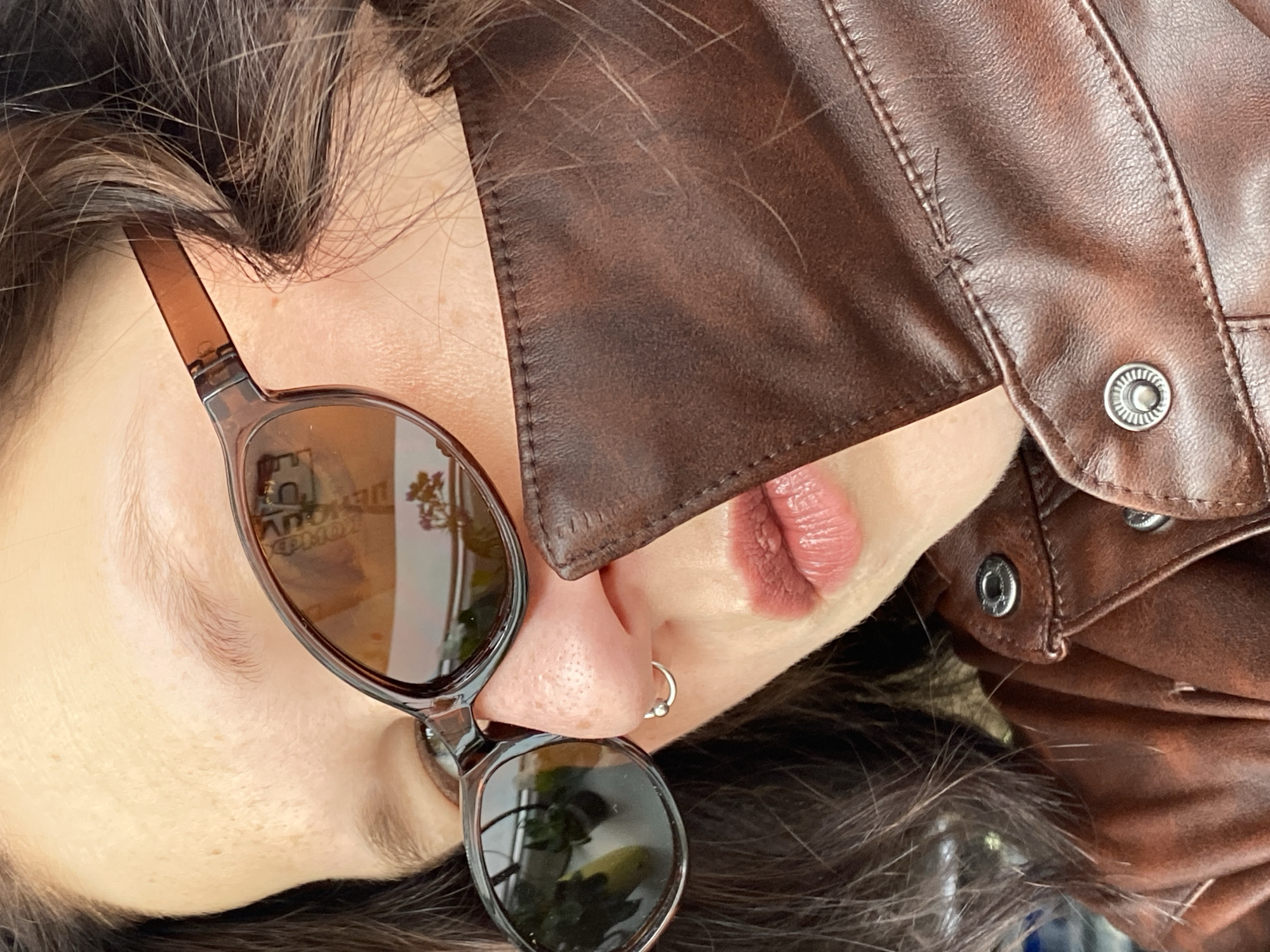 The image is a rotated, close-up portrait featuring the side profile of a person, likely taken in Chaikovsky, Russia. The individual's face is positioned so their head is tilted to the right, showing parts of their left cheek, nose, lips, and hair.
The main subject is a person with fair skin, dark brown hair that is partially visible around their face and resting on their shoulder and a brown leather (or faux leather) jacket. They are wearing sunglasses with translucent brown frames and dark tinted lenses, which reflect some indistinct foliage or outdoor scenery. A small, silver hoop nose piercing with a bead is visible on their left nostril. Their lips are painted with a matte, dusty rose or nude pink lipstick. A small bump is visible on the upper lip.
The person is wearing a brown jacket with visible stitching and metal snap buttons on the collar and front flap. The jacket appears to have a slightly distressed or textured finish. Strands of the person's dark brown hair are visible scattered across the jacket's surface in certain areas.
The overall activity is a posed portrait or selfie, focusing on fashion and close-up details. The lighting suggests daytime, possibly outdoors or near a window, given the reflections in the sunglasses. The emotion is not clearly conveyed due to the partial view of the face, but the image has a casual and stylish aesthetic. Blurred, mirrored text, possibly "D KOWAU" or similar characters, is faintly visible in the reflection of the sunglass lenses but is unreadable.