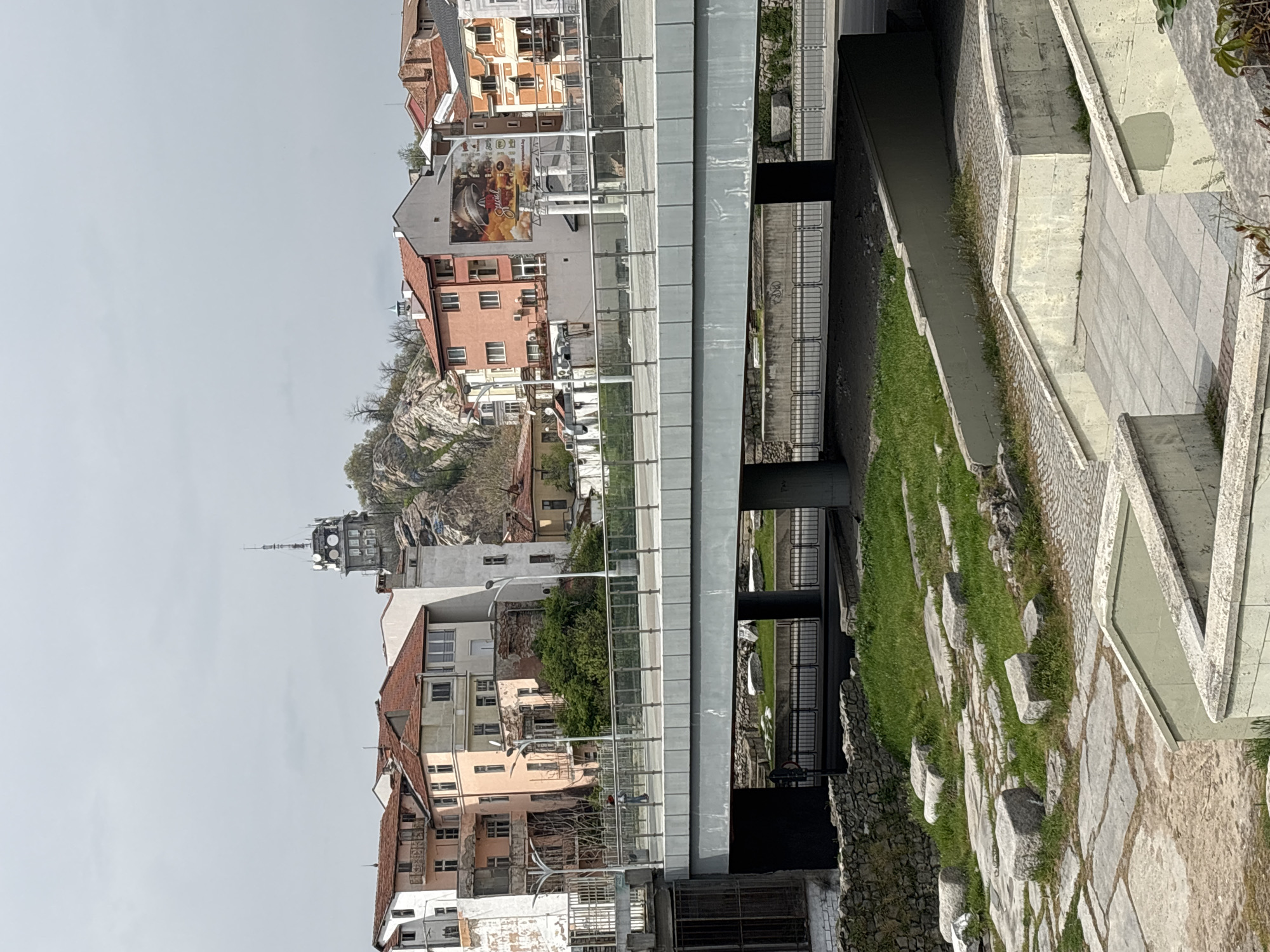 This is an eye-level, landscape-orientation photograph of a cityscape in Plovdiv, Bulgaria. The buildings vary in color and style, including peach, orange, grey, and white. A tall tower with communication equipment on top stands on a hill in the background. A modern metal and glass bridge spans a small concrete-lined waterway with grassy banks. The foreground includes a stone path and concrete structures with geometric shapes. The sky is overcast, providing a neutral grey background.