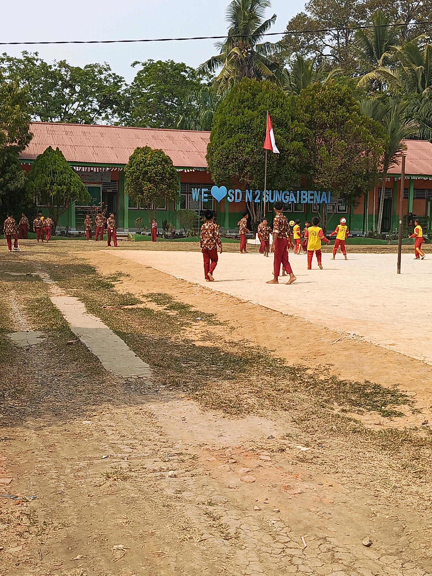 The image depicts an outdoor scene at a school in Megang Sakti IV, Indonesia, likely during daylight hours given the bright lighting and clear sky. In the background, a school building with a red roof and orange and green walls is visible. A large sign with blue lettering and a heart symbol reads "WE ♡ SD N 2 SUNGAI BENAI". In front of the building, a group of children are engaged in various activities. Some children are standing, while others appear to be playing soccer in a dirt field. Many of the children are wearing matching outfits, predominantly red and white with patterned shirts, suggesting a school event or uniform. A flagpole with an Indonesian flag is also present. The foreground shows a dirt and grass area, indicating a schoolyard or playing field. The overall environment appears to be a tropical or sub-tropical setting, with numerous trees and lush green foliage surrounding the school.
