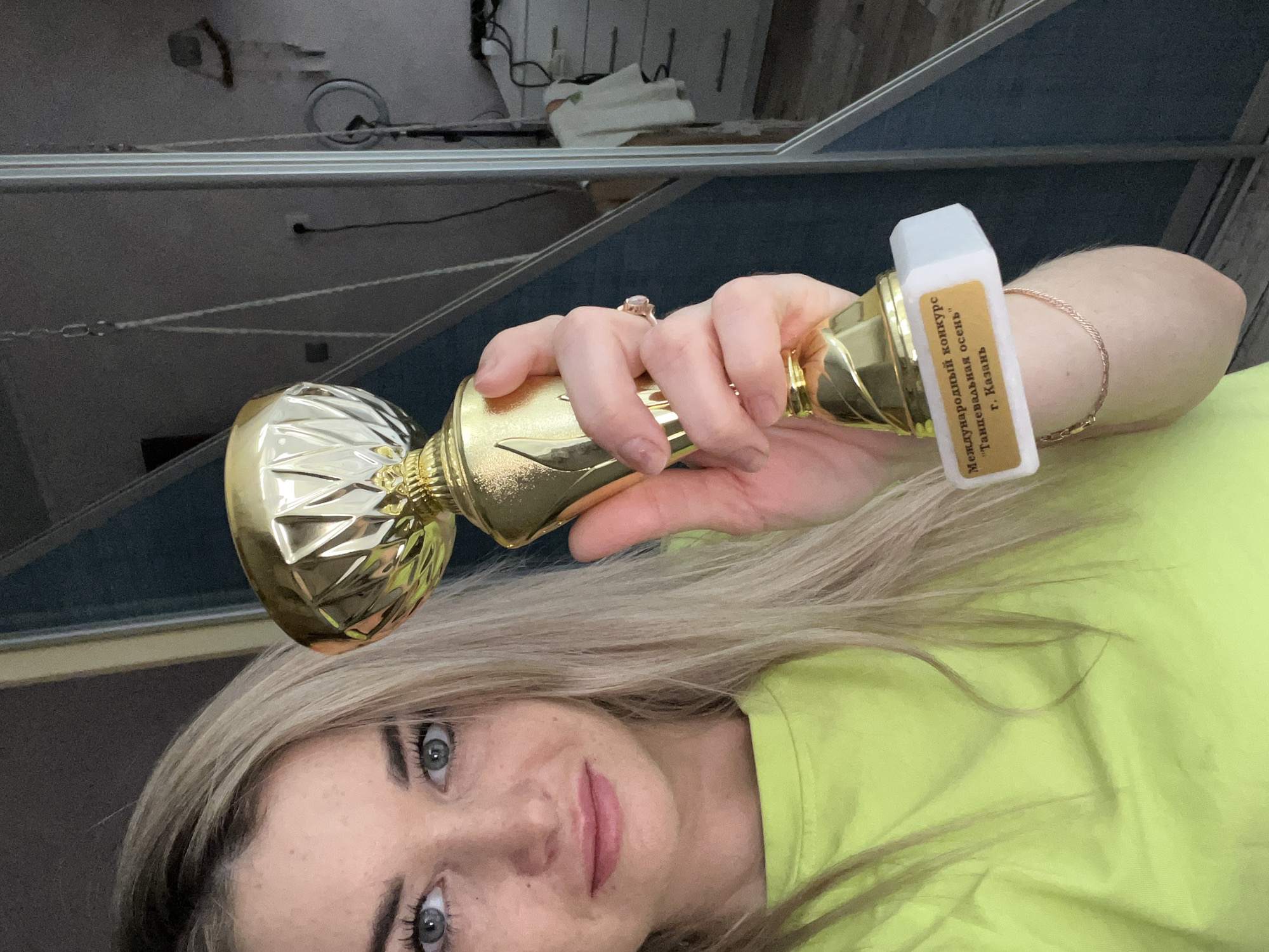 A woman with blonde hair and blue eyes is holding a gold-colored trophy. The trophy has a white label on the base with Russian text that includes "Международный конкурс Таиппевальнвя осень г. Казань", which translates to "International Contest of Taippevalny Autumn, Kazan City". The woman is wearing a lime green shirt and a ring on her left hand. She also has a gold bracelet. The background is blurry and appears to be an indoor setting, possibly a room with a shelf or mirror. There is not enough information to determine that this scene is in the city of Chaikovsky, Russia.