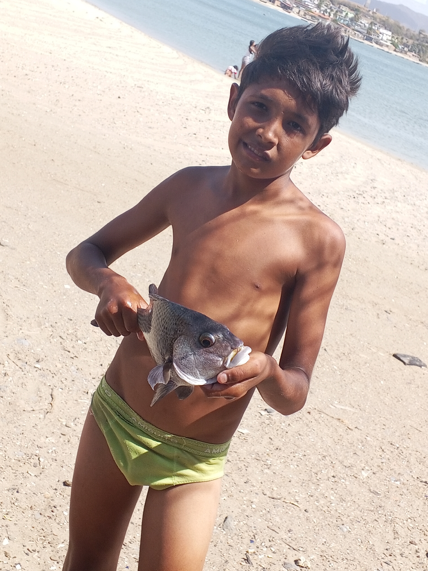 A shirtless young boy stands on a sandy beach, holding a fish in his hands. He is facing the viewer, smiling slightly. The background shows a beach with other people in the distance and a body of water. In the distance, there are buildings and hills which might indicate the presence of the city of Juan Griego, Venezuela. The boy is wearing a light green swimsuit. The fish has a dark gray color with white areas near its mouth.