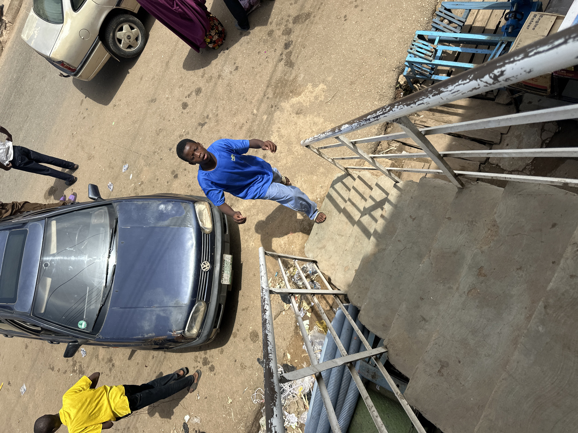 A medium shot, high-angle view captures a lively scene in the city of Jos, Nigeria. In the foreground, a young boy, dressed in a bright blue t-shirt and light denim jeans, with sandals on his feet, is in mid-stride, looking towards the viewer with wide eyes and an open mouth. He appears to be on a concrete staircase, which is lined with a weathered metal railing. To the left of the stairs, stacks of gray plastic pipes are visible.
In the background, a dark blue Volkswagen sedan is parked on a dusty, sun-drenched road. A person wearing a yellow t-shirt and black trousers is standing near the car, facing away from the viewer. Further down the road, another vehicle, a light-colored sedan, is parked. A person in a purple garment is partially visible to the right, near the light-colored car. The environment suggests a busy urban street with some debris and scattered pieces of paper on the ground. The overall impression is one of everyday life unfolding in a Nigerian city.