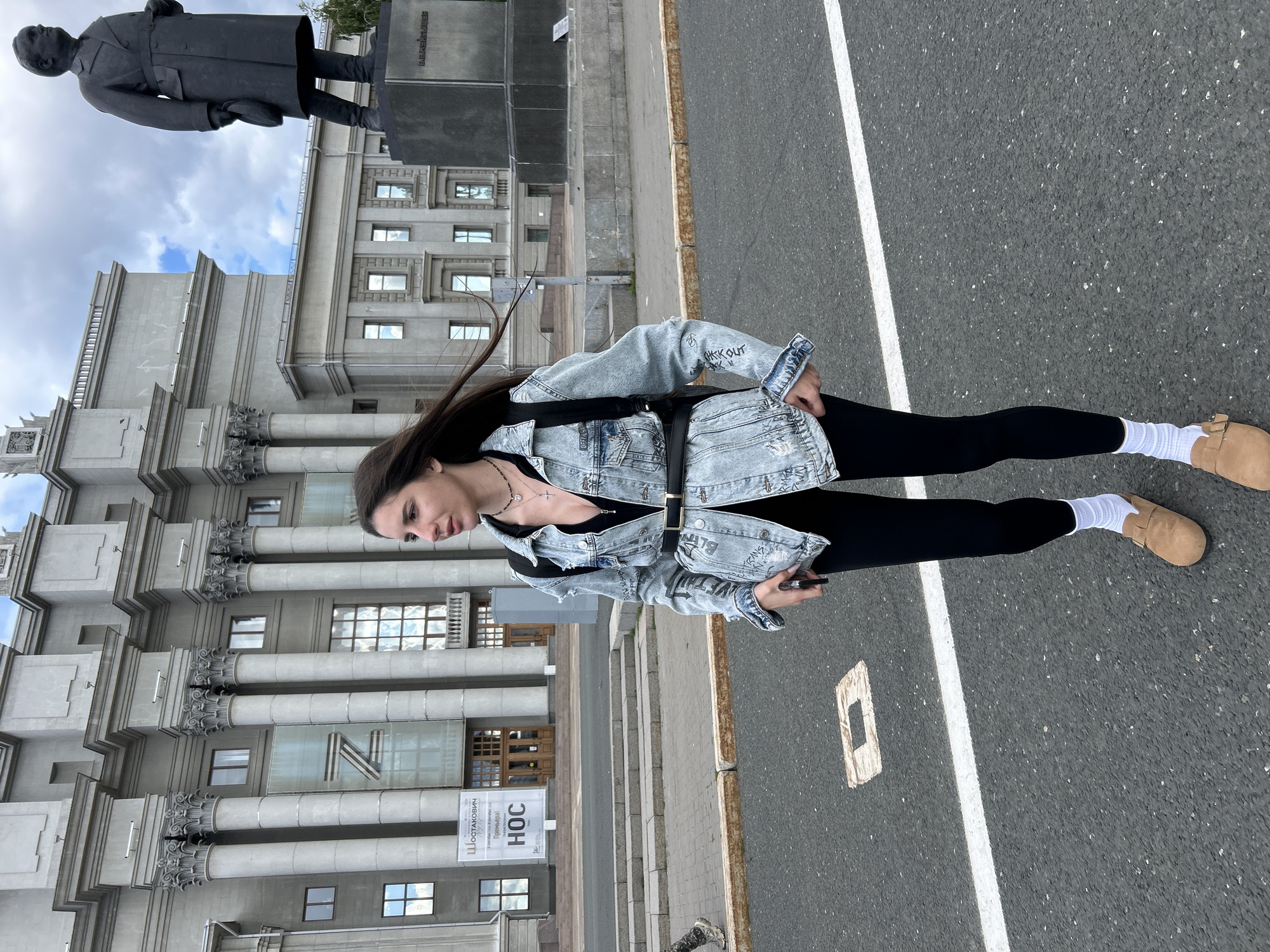 A young woman with long dark hair, wearing a denim jacket, black top, black leggings, white socks, and tan clogs, stands on a paved street in front of a large, gray, classical-style building. A statue of a man in a coat and hat stands to the right of the building. The building has large columns and windows. A large, translucent banner with the letter "Z" is visible on the building's facade, along with text that appears to be in Cyrillic. The woman is holding a dark-colored smartphone in her right hand and looking to her left. The sky is overcast with some blue patches. The location is the Novokuybyshevsk Oil Refinery Palace of Culture in Novokuybyshevsk, Russia.