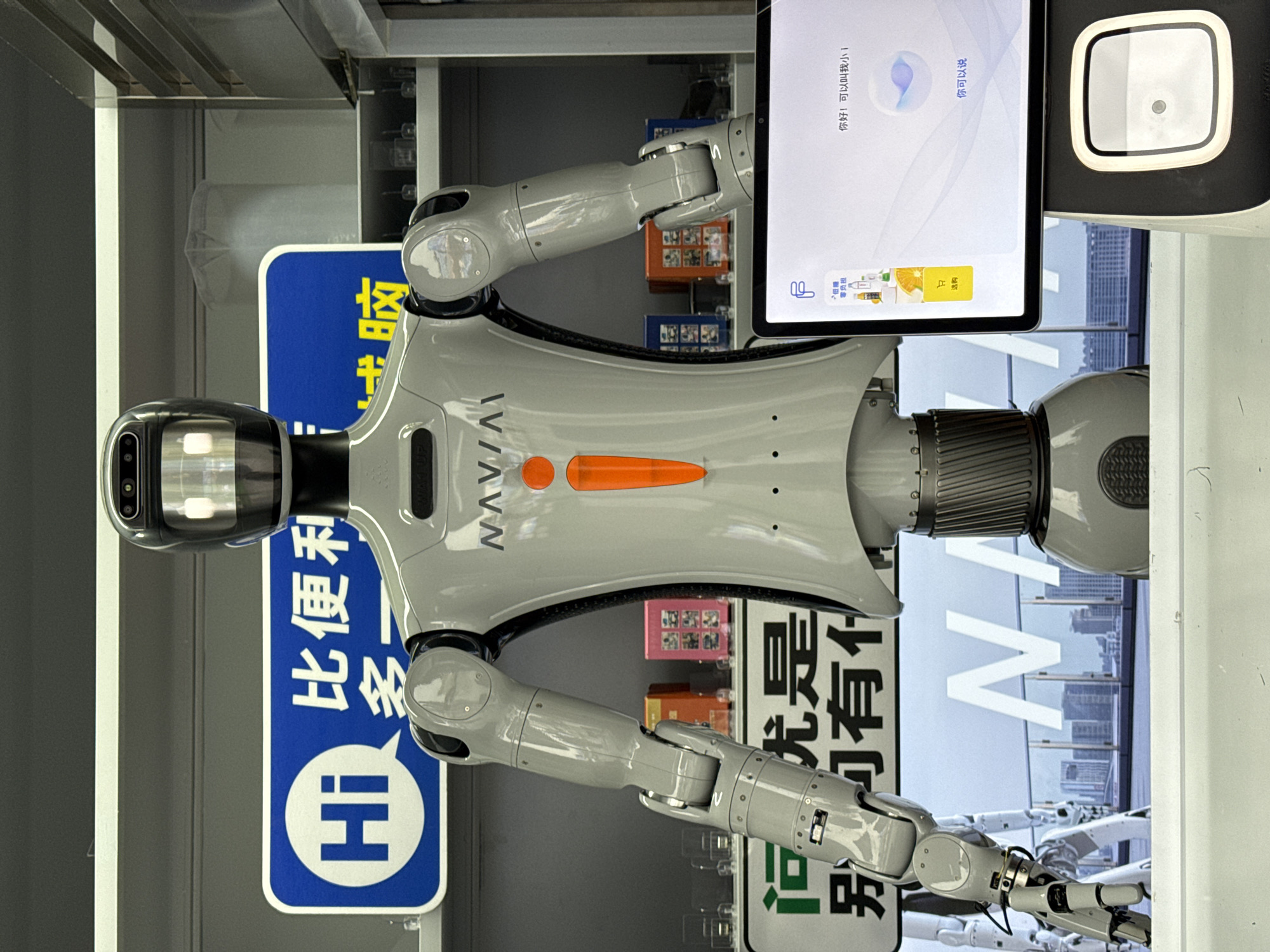 An indoor shot depicts a full-body, light gray humanoid robot with a dark gray, rounded head. The robot has a prominent orange exclamation point symbol on its chest and "NAVIAI" written above it. Its arms are articulated and appear to be made of multiple segments. In front of the robot is a tablet device displaying a user interface with Chinese text and graphics, including a shopping cart icon and a greeting that reads "你好!可以叫我小 i" (Hello! You can call me Xiao i). Below the tablet is a black base with a square scanning area.
Behind the robot, a blue sign with white text says "Hi" in a speech bubble, followed by Chinese characters. To the left of this sign, another green sign with white Chinese text is partially visible, reading "河边是... 别有什麼" (Riverside is... what else). Shelves behind the robot display small, colorful boxes and some clear containers. In the background, a large screen shows a cityscape with tall buildings, suggesting an urban environment. The scene takes place in what appears to be a commercial or retail setting, possibly a convenience store or a demonstration area. The overall lighting is bright and even. The location is Nanshan District, China.