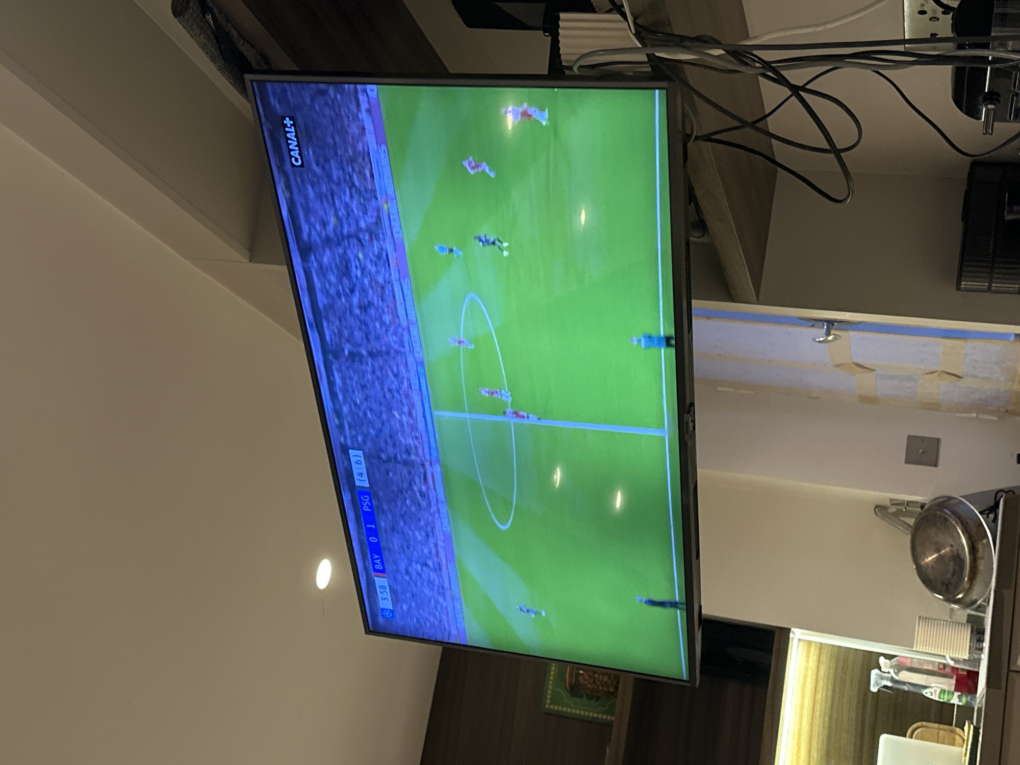 The image shows a television screen displaying a soccer game, specifically a match between Bayern Munich (BAY) and Paris Saint-Germain (PSG). The score is BAY 0-1 PSG, and the time is 3:58. The channel logo "CANAL+" is visible in the top left corner. The television is located in a kitchen area with cabinets, appliances, and a window. The location is suggested to be in Neuilly-sur-Seine, France, based on the French TV channel. There are no people clearly visible in the frame.