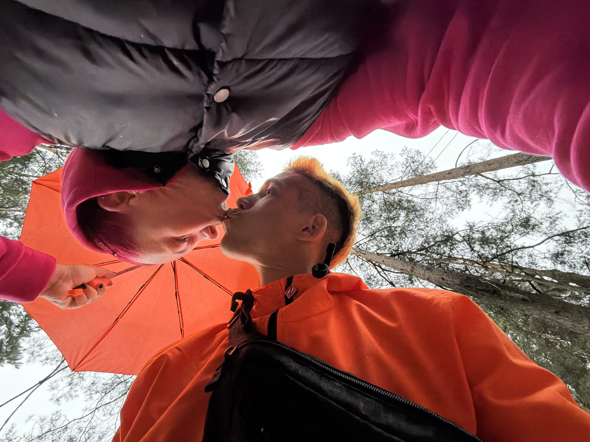 In this low-angle, close-up shot, two individuals are embracing and kissing under a bright orange umbrella. The person on the left is wearing a pink hooded jacket over a dark puffer coat with white snaps. Their head is tilted back, with pink-dyed hair visible. The person on the right is wearing an orange jacket with a black backpack strap visible. They have spiky, orange-dyed hair. Both individuals' faces are obscured by their kiss. The background shows trees and a cloudy sky, suggesting an outdoor setting, possibly in the city of Chaikovsky, Russia. The overall impression is one of intimacy and affection.