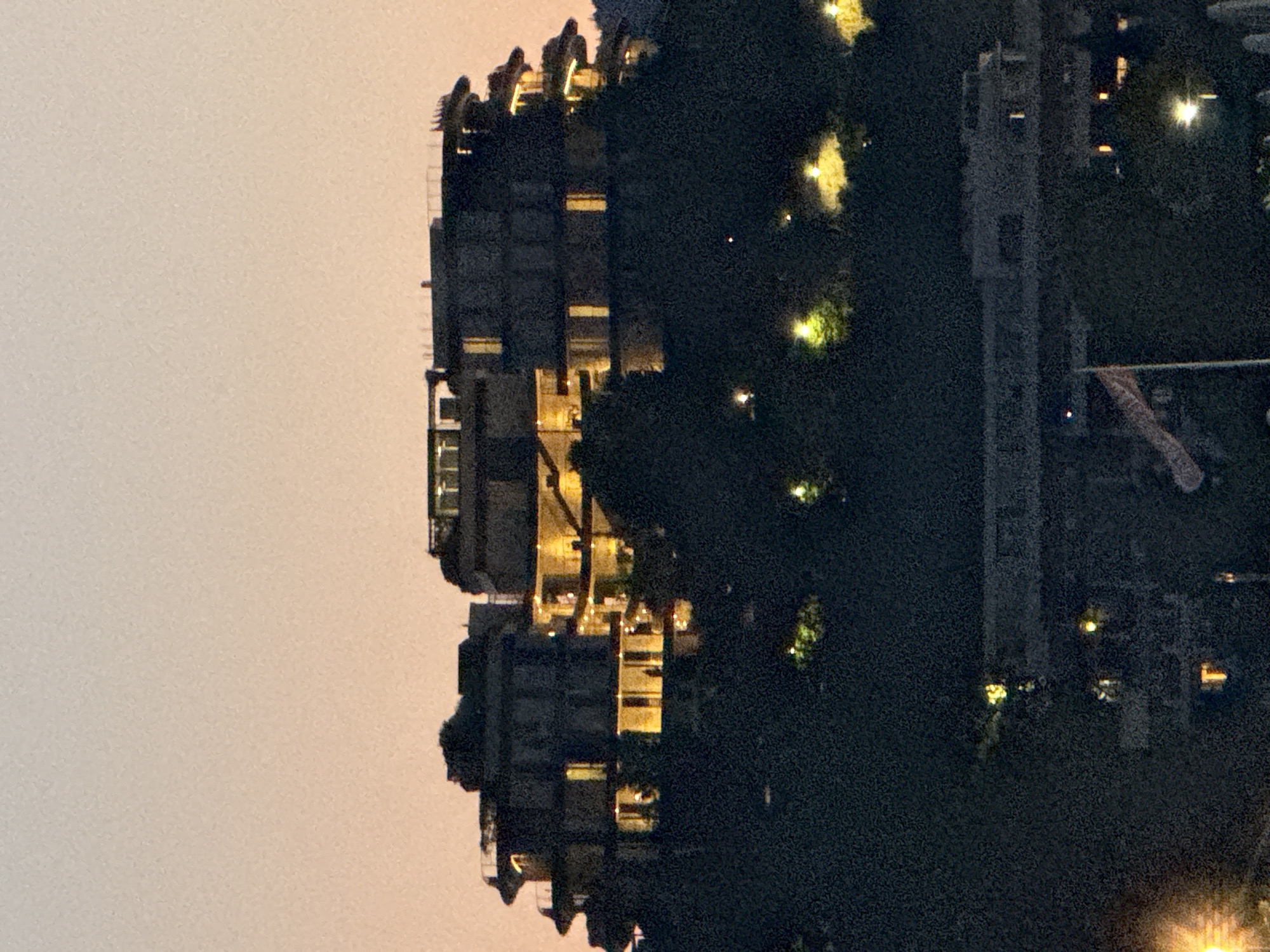 The image depicts a multi-story building on a hill overlooking a body of water, likely the Bay of Kotor, in Boreti, Montenegro. The sky is a gradient of pale orange to light pink, indicating dusk or dawn. The building itself appears to be a modern hotel or residential complex, with numerous windows illuminated by warm, yellow light. The structure has curved balconies and a tiered design. Below the building, dense, dark foliage obscures the lower parts of the hill and the foreground. Scattered lights are visible within the trees, suggesting other structures or streetlights. In the lower portion of the image, a long, horizontal, lighter-colored structure, possibly a ferry or a train, is partially visible. A flagpole with a triangular flag is also present in the lower left quadrant of the image. The overall impression is one of a serene evening scene in a coastal town.