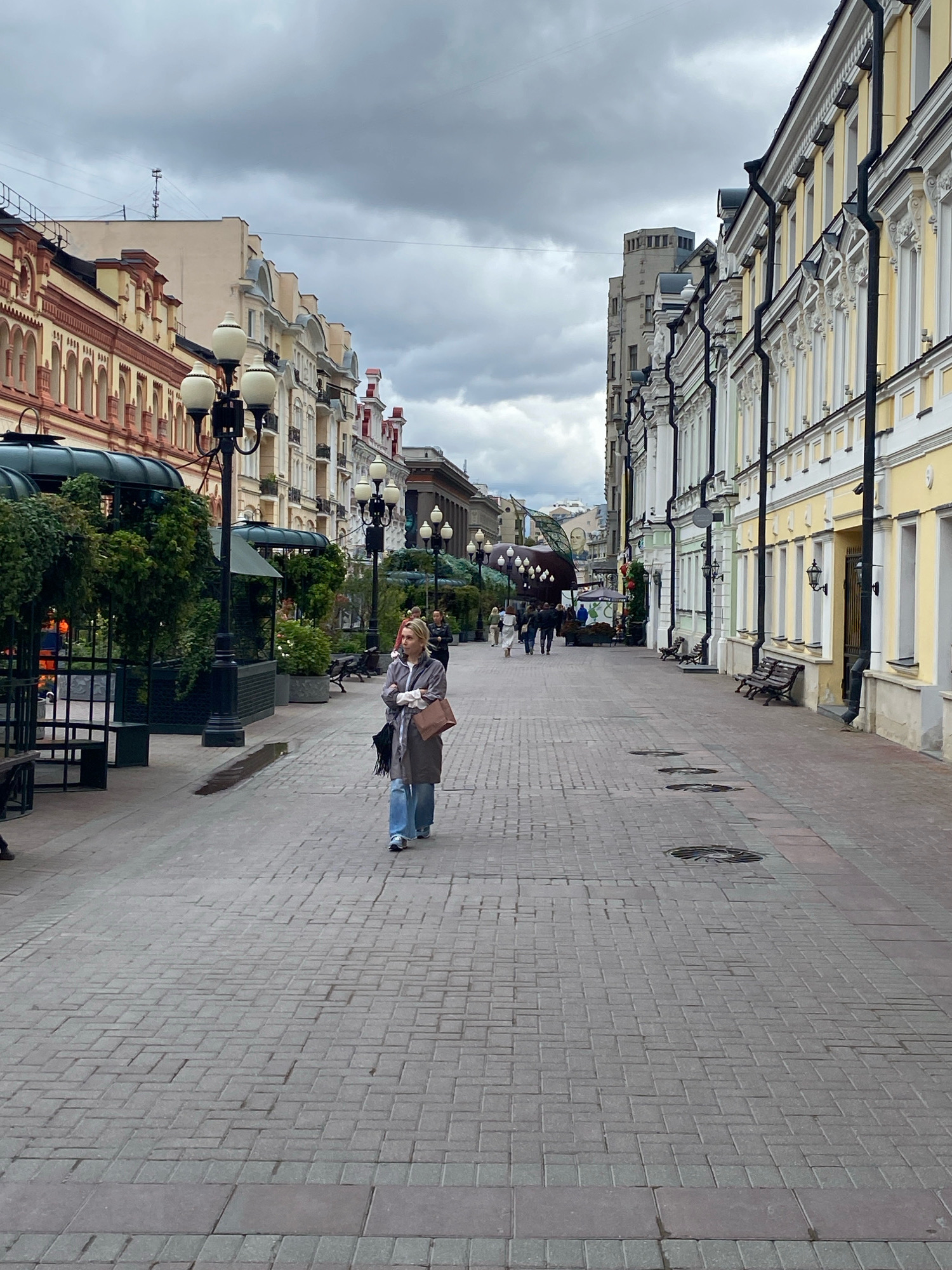 The image depicts a pedestrian street in Moscow, Russia, under an overcast sky. The street is paved with gray and darker red bricks, and lined with buildings on both sides.
On the left side of the street, the buildings feature ornate facades with red and yellow coloring, arched windows, and decorative architectural elements. There are trees and greenery within fenced enclosures, as well as street lamps and benches.
On the right side of the street, the buildings are predominantly yellow with white decorative trim, and also feature numerous windows. Black downspouts are visible along the building facades. Several street lamps and benches are also present on this side.
A single woman is walking towards the viewer in the center of the street. She is wearing a gray coat, blue jeans, and carrying a brown bag. In the background, other pedestrians can be seen walking in both directions, and there are also some people seated at outdoor tables, likely at cafes or restaurants. The overall impression is of a commercial or entertainment district.