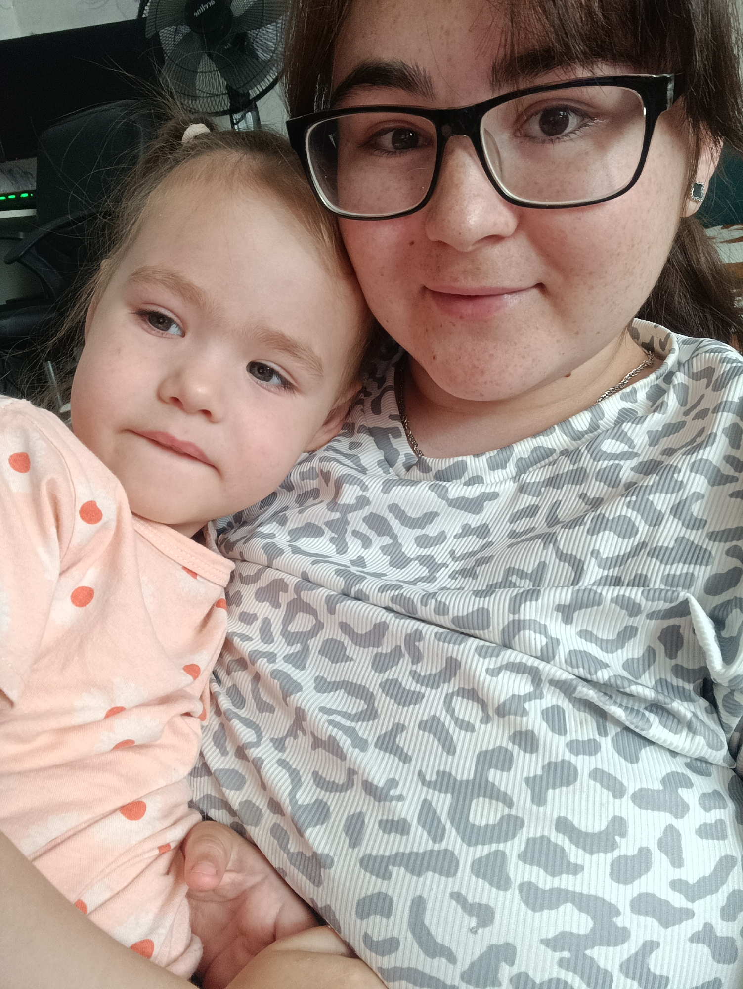 This is a selfie taken by a woman wearing glasses and a grey and white leopard-print shirt. She is holding a young child, who is wearing a light peach-colored shirt with orange polka dots. The child is leaning on the woman's shoulder. The background is blurry but appears to be indoors, with a fan visible behind them. The image does not contain any information about the location in the city of Noviy, Russia.