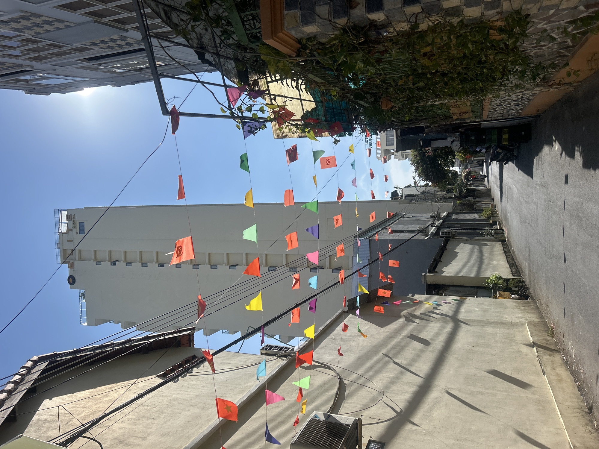 The photo is taken in Bac Nha Trang Ward, Vietnam. It shows an alleyway decorated with strings of colorful flags hanging between buildings. The sky is visible above, with a bright sun. A tall, white building is seen in the background. The alleyway has a paved surface, and the buildings on either side are a mix of residential and commercial structures. Some buildings have tiled roofs, while others have flat roofs. Shadows from the flags and buildings are visible on the alleyway floor. There is no presence of people in the alley.