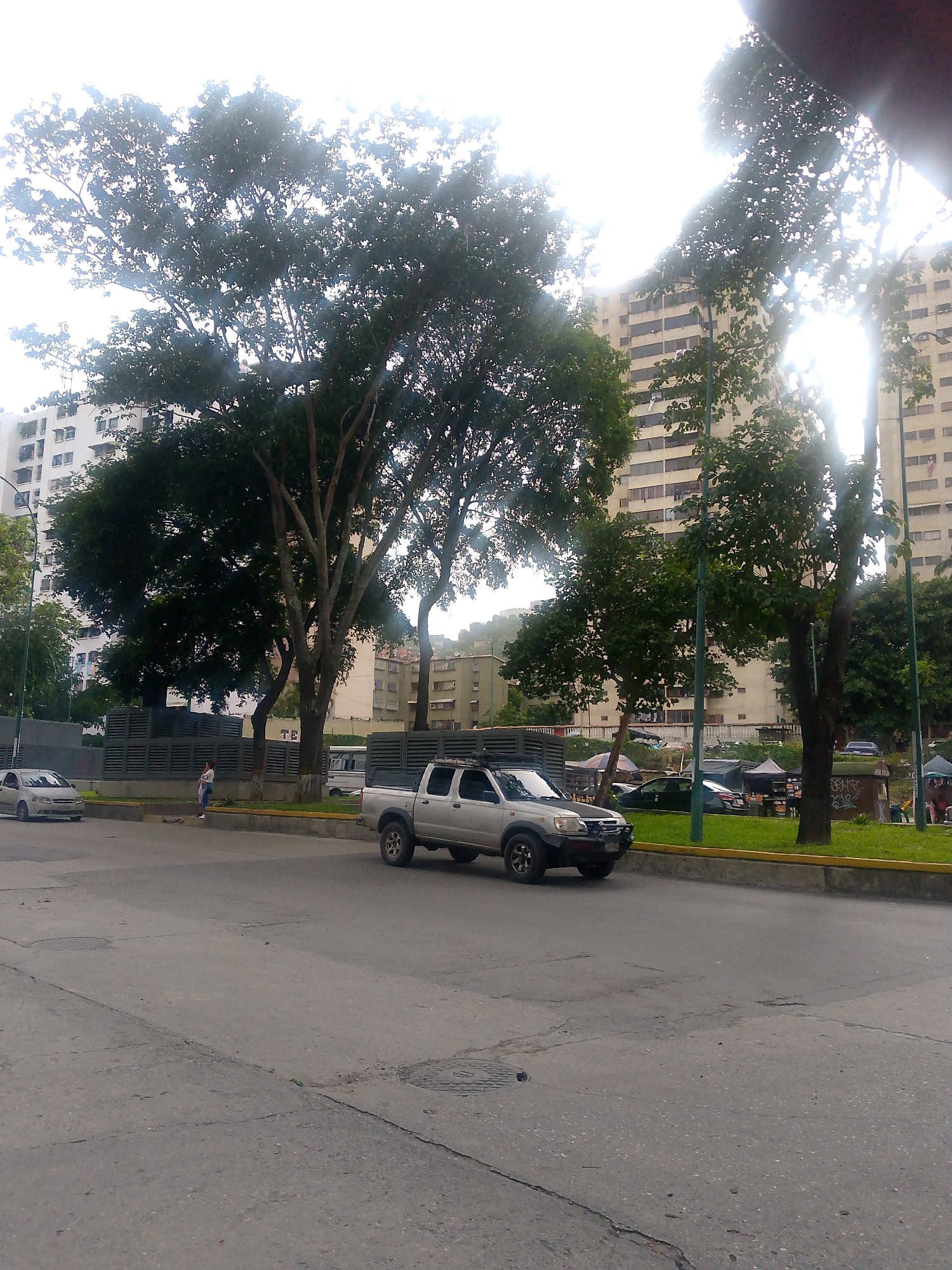The image depicts an outdoor urban scene, likely in Caracas, Venezuela, under a bright, overcast sky. In the foreground, a paved road shows signs of wear with cracks and manhole covers. A silver pickup truck is driving on the road, facing left. To the left of the truck, a silver sedan is parked on the side of the road. A woman stands on the sidewalk near the sedan, facing the truck.
Behind the road and sidewalk is a gray fence with a slatted design. Beyond the fence, a lush green area with trees and grass is visible. In the background, tall, multi-story apartment buildings with numerous windows line the skyline. The buildings appear to be modern residential structures. The trees in the scene are large and leafy, with substantial trunks, suggesting a mature urban landscape. The overall impression is a typical street view in a densely populated city.