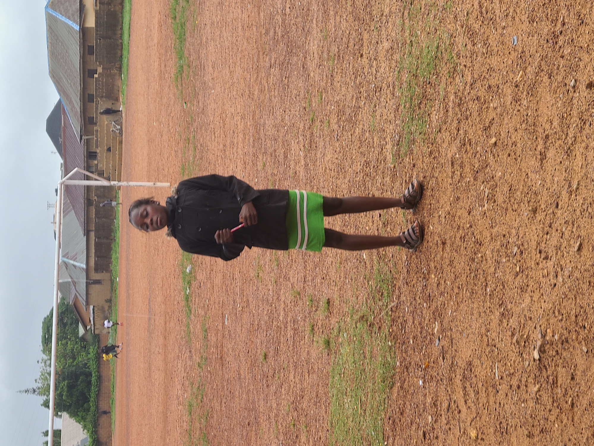 A young person stands on a large, reddish-brown dirt field with scattered patches of green grass. The individual, who appears to be a child or young adolescent, is positioned slightly to the right of the center, looking directly at the camera with a neutral expression. They are wearing a dark, possibly black, long-sleeved jacket over bright green shorts with white stripes, and dark sandals. In their hands, they hold a slender pink object.
In the background, to the left, are several buildings with varying roof types, including corrugated metal and darker structures, suggesting residential or community buildings. A white goalpost frame is visible in the middle-ground on the left. Further back, near the buildings, a person is seen standing next to a white bicycle. More distant figures are visible on the field, appearing to be walking or playing. Green trees and foliage are present along the far left edge of the scene. The sky is overcast, indicating cloudy weather during daylight hours. The scene depicts an open, outdoor area, likely a recreational field or community space in a rural or semi-urban setting. No visible text is present in the image.