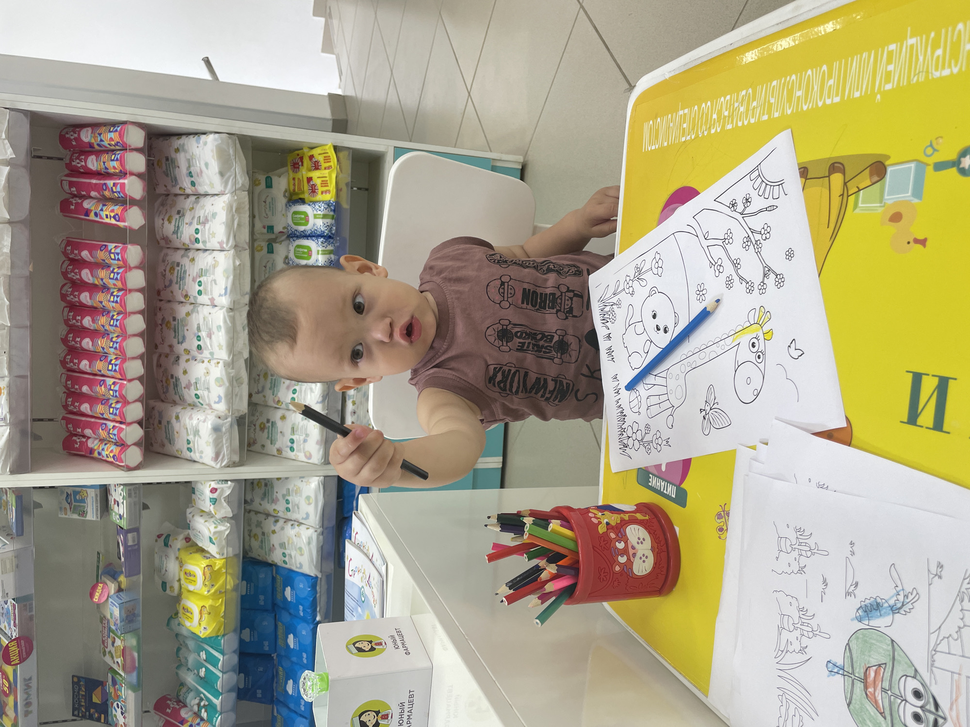In a bright, indoor environment that appears to be a pharmacy, a young child with light brown hair is seated at a yellow table, engaged in coloring. The child is wearing a brown t-shirt with a graphic print. The table is adorned with coloring pages, one depicting a giraffe and another featuring cartoon characters. A red cup filled with colored pencils stands on the table, and a blue pencil is positioned on one of the coloring pages. In the background, shelves are stocked with various products, including diapers and other baby care items. The child is holding a broken black pencil in their right hand. The scene suggests a creative activity for children within a retail setting. The text "ЮНЫЙ ФАРМАЦЕВТ" (Young Pharmacist) is visible on a white box near the child. The overall impression is one of a child-friendly space within a commercial establishment. Based on the Cyrillic text and typical pharmacy product packaging, the location is likely in Russia, and given the specific branding and product types, it could be in the city of Perm, Russia.