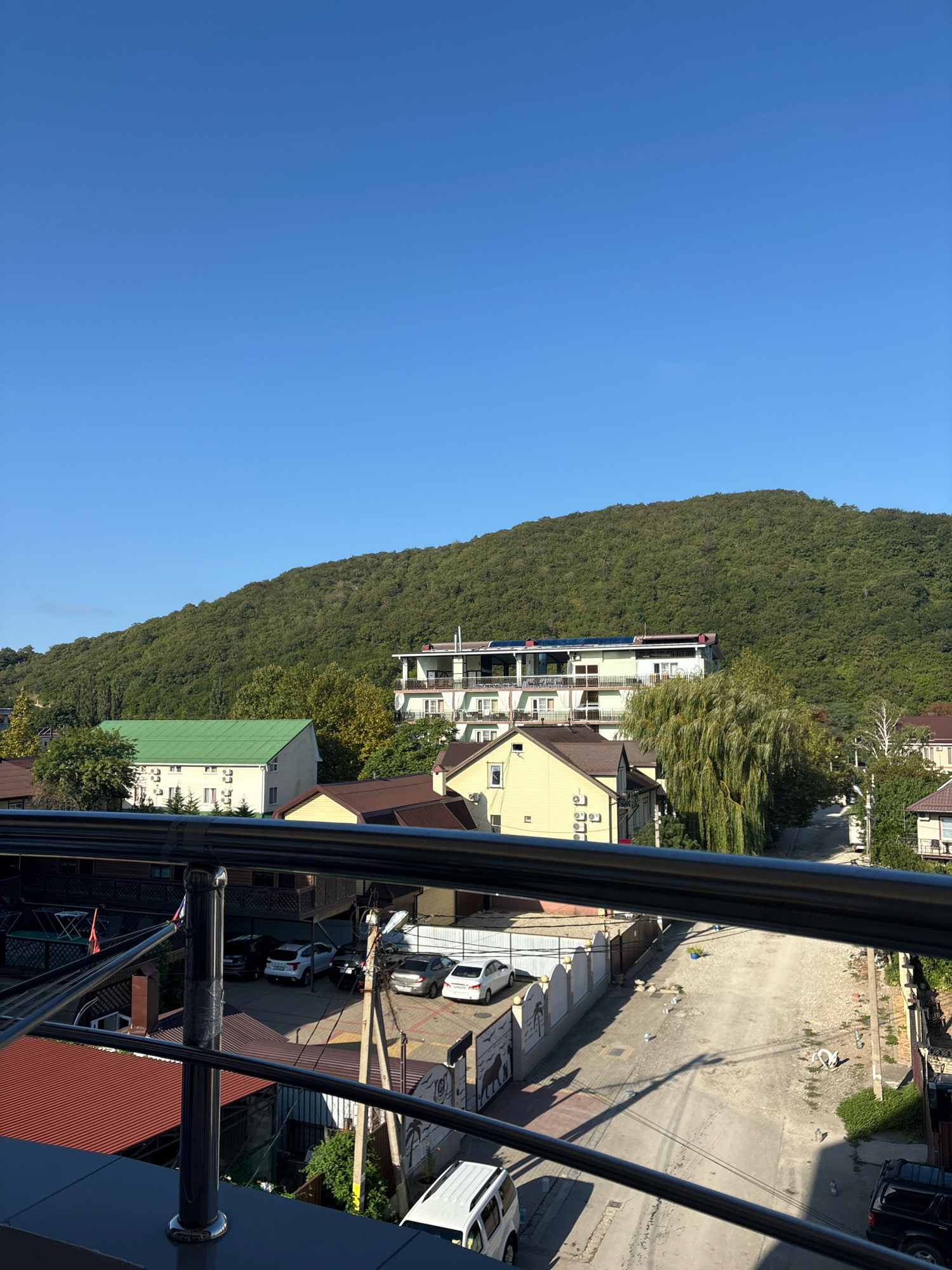 This is a daytime, eye-level, outdoor shot of a rural street scene in Arkhipo-Osipovka, Russia. In the foreground, a dark metal railing with black tape wrapping around a support pole is visible. Behind the railing, a gravel road runs through a residential area. Several parked cars, including white sedans and a black SUV, are present on the street and in adjacent parking areas. Buildings with light-colored facades and brown or green roofs line the road, with some featuring air conditioning units. In the background, a dense, green, tree-covered hill rises under a clear blue sky.