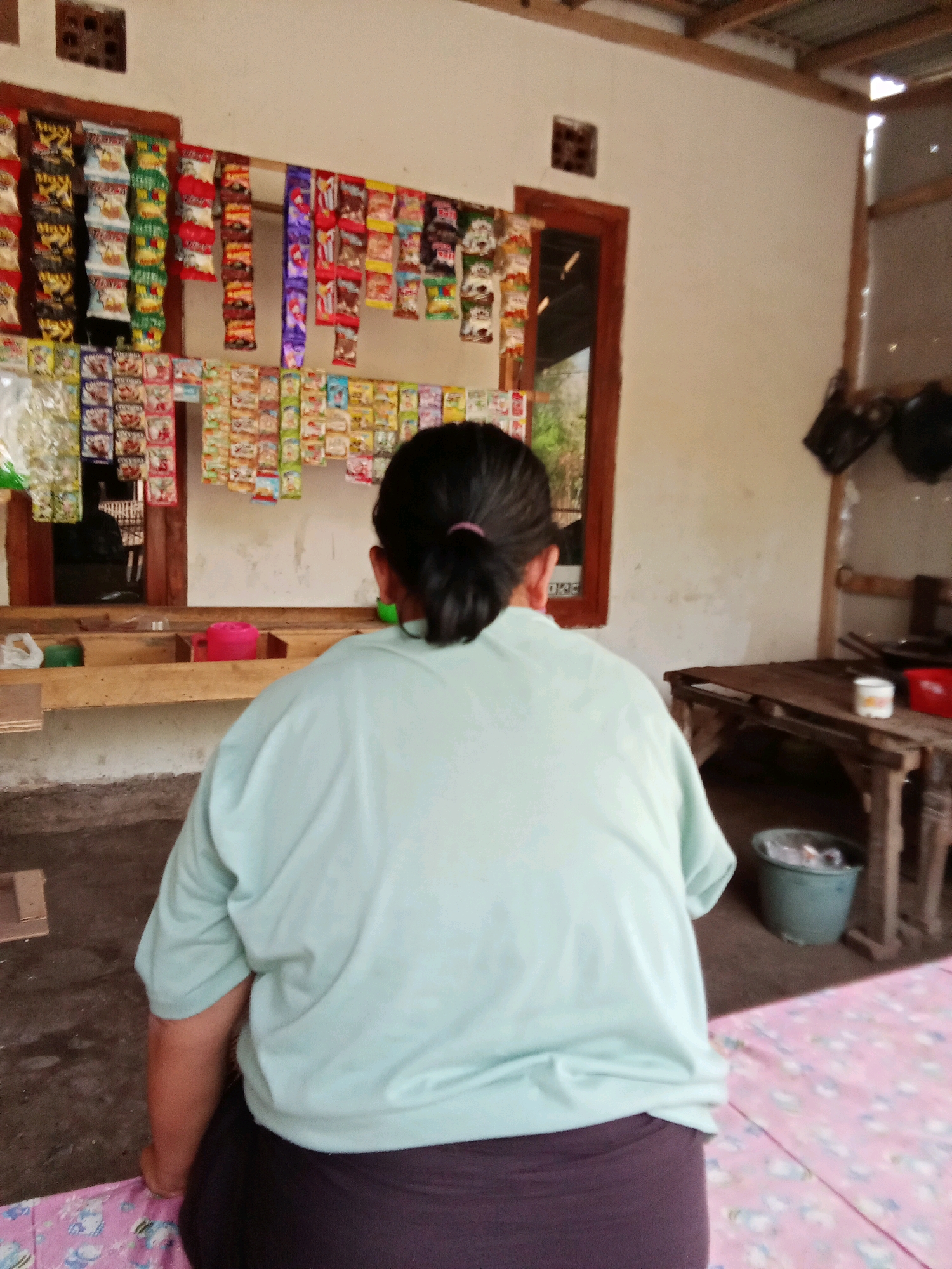 The image depicts a rear view of a person wearing a light blue t-shirt and dark pants, positioned indoors. They are facing a display of small packaged goods, likely snacks or candies, hanging on a wall above a wooden shelf. The environment appears to be a small shop or warung. In the background, there is a wooden table with various items, including a white cup and a red container, and a green bucket on the floor. The architecture suggests a rustic setting, possibly in Selowogo, Indonesia, with unfinished wooden beams and a corrugated metal roof visible at the top. The lighting indicates an indoor setting with natural light entering from an opening on the right side of the image.
