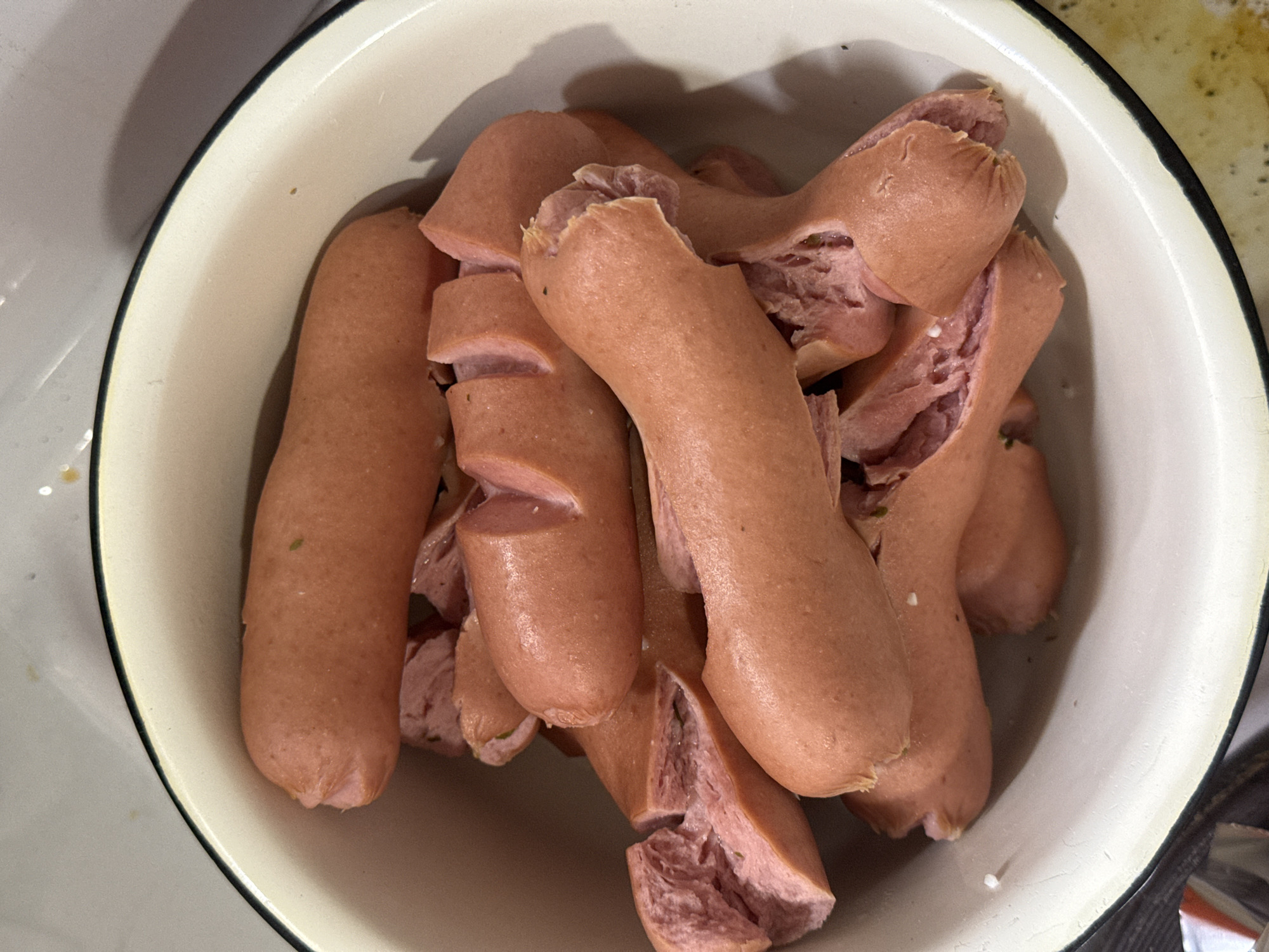 An overhead shot shows a white enamel bowl with a black rim filled with cooked, cut sausages. Some sausages are whole, while others are cut into segments with vertical slits. The sausages are pale pink and appear to be plump. The location and interaction are not identifiable from this image.