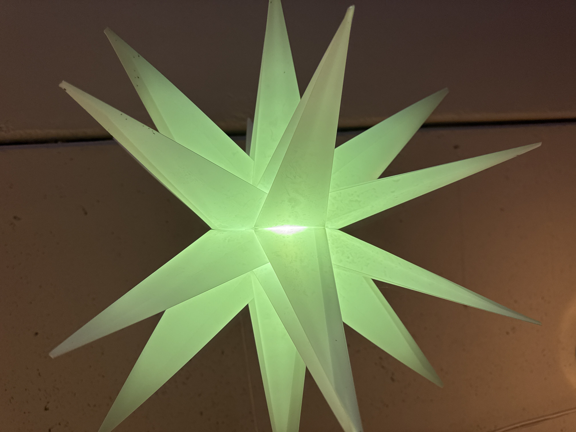 An illuminated, pale green, 3D star-shaped ornament is suspended against a dark, textured ceiling. The ornament has numerous pointed tips radiating outwards, creating a starburst effect. A bright light source emanates from the center of the ornament. The background appears to be a ceiling with some minor imperfections. No people or actions are depicted. The location is not identifiable from the image.