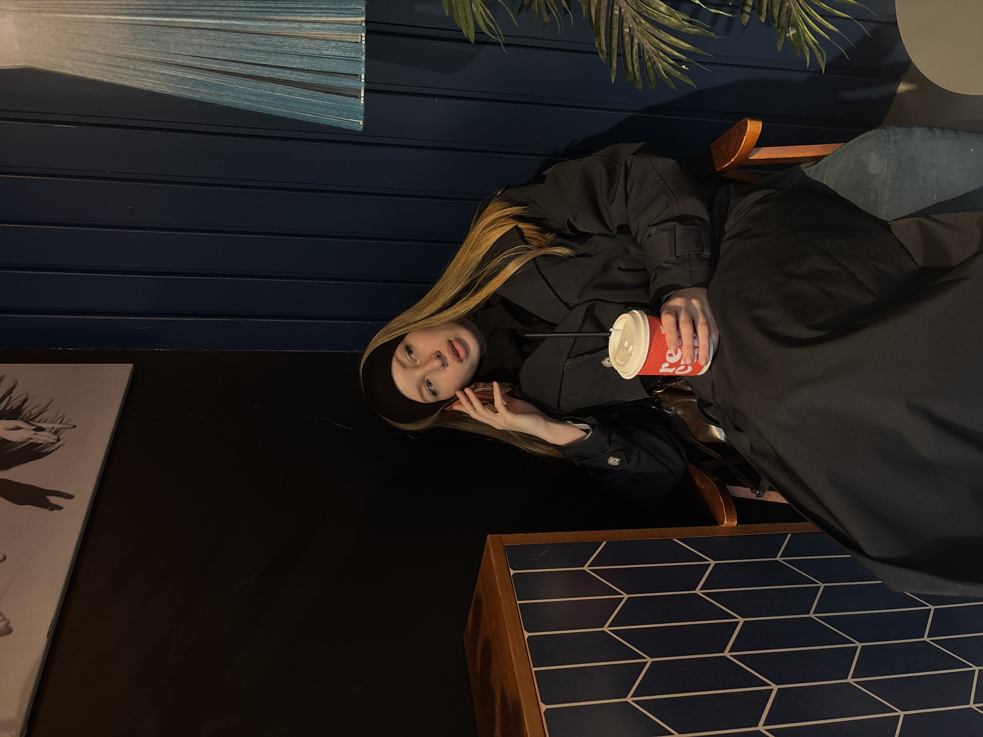 A young woman is seated indoors, likely in a cafe or lounge in Perm, Russia. She has long, straight blonde hair and is wearing a black top or jacket with a black headband. She looks towards the camera with a casual, slightly playful expression, her tongue subtly extended.
She holds a red and white paper cup with a white lid and a black straw in her right hand; the word "COFFEE" is partially visible in white lettering on the cup. Her left hand is near her jawline. The background features dark blue wooden paneling. To her upper left, blue slatted blinds or a decorative panel are visible, catching some light. Artificial palm-like leaves are in the upper right. A table with a dark blue geometric pattern of parallelograms/hexagons outlined in white and wooden edges is in the lower right foreground. Part of a framed black and white artwork is visible on the far left. The lighting is dim, suggesting an evening or interior setting.