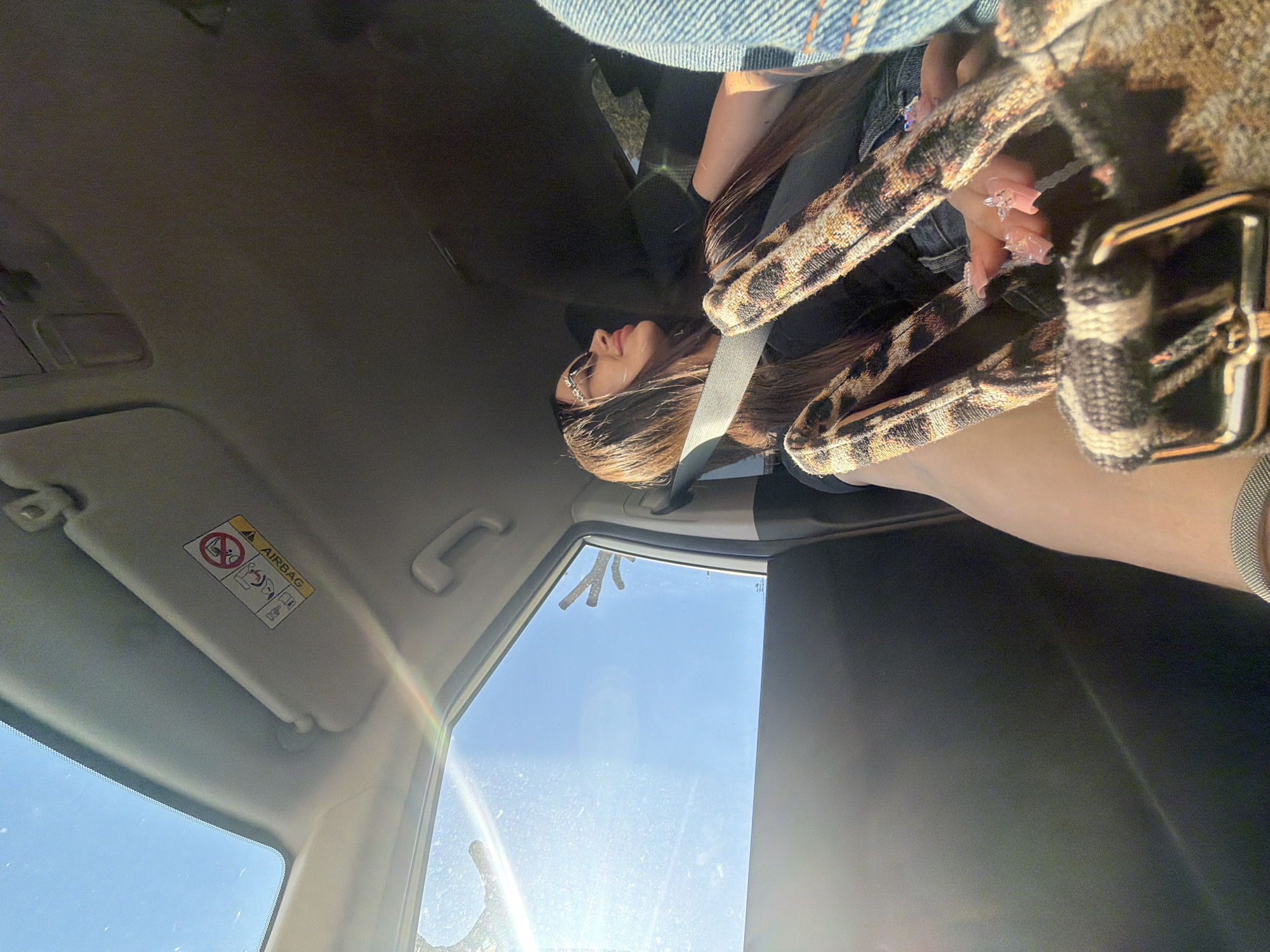 The image, taken from inside a car, shows a woman seated in the passenger seat during what appears to be a bright, sunny day. She has long, dark brown hair and is wearing sunglasses, a black top, and a denim jacket, with a seatbelt diagonally across her chest. Her face is visible in profile, looking slightly upwards and to the right. Her hands, adorned with long, pink, decorated fingernails, are holding a bag or strap with a distinctive leopard print pattern.
The car's interior is visible, including the light-colored ceiling and a sun visor on the passenger side. An "AIRBAG" warning label with pictograms for child and pregnant passenger safety is clearly displayed on the sun visor. Through the car's windshield and side window, a clear blue sky is visible, along with sunlight causing lens flares and reflections. Some branches or bare trees can be seen through the side window against the sky.
The scene suggests the woman is a passenger in a vehicle, either stationary or in motion, on a clear day in Izhevsk, Russia. The bright sunlight and blue sky indicate daytime. Her posture appears relaxed, and the overall impression is peaceful.
