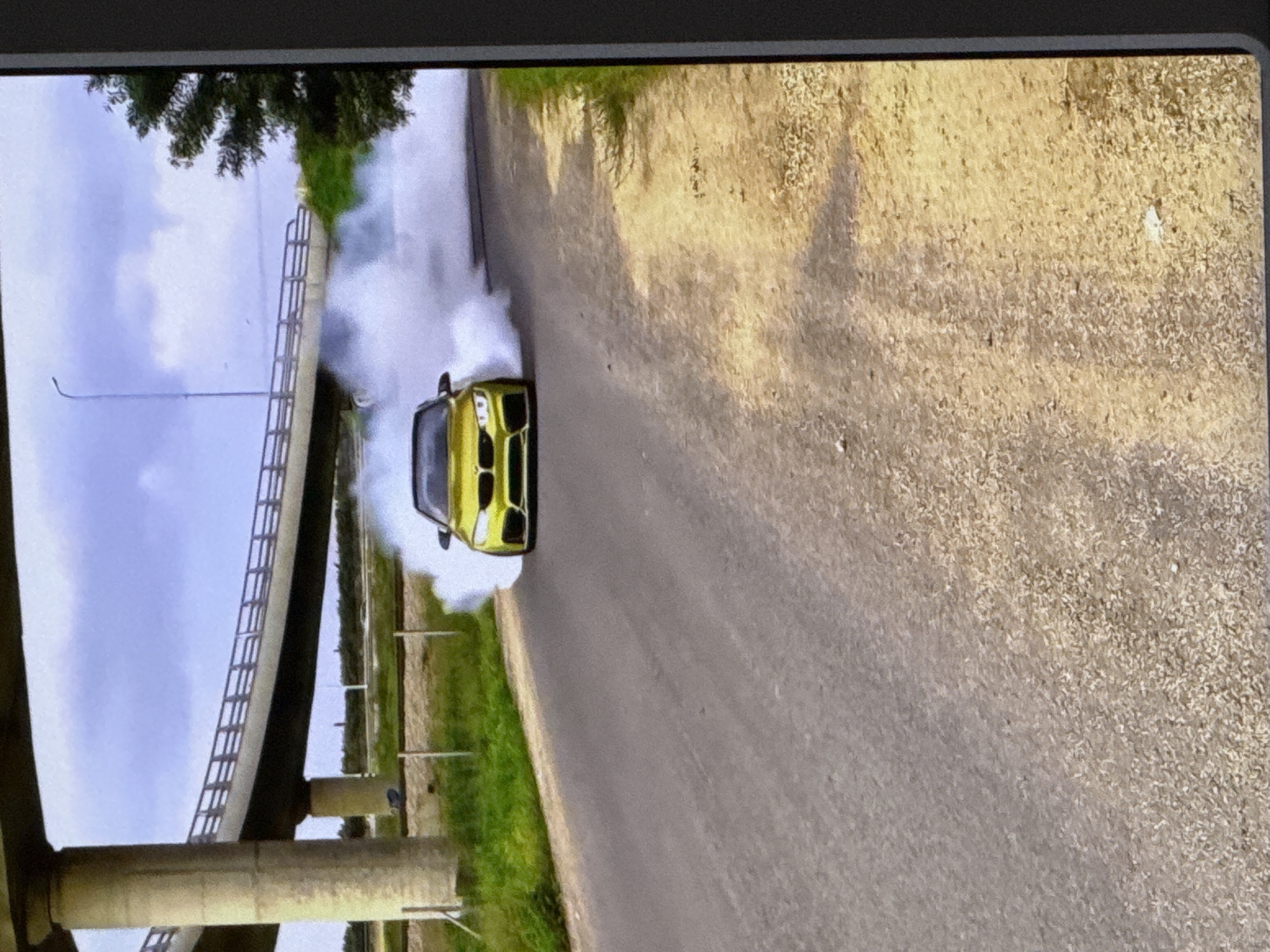 A yellow BMW M4 is captured mid-drift, emitting a large cloud of white smoke on a paved road. The car is facing the camera, with its headlights on. The scene appears to be on the outskirts of a city, with a large overpass or bridge structure in the background. The surrounding environment includes dry, grassy terrain and a dirt shoulder. The sky is overcast with clouds. The location is identified as Cahul, Moldova.
