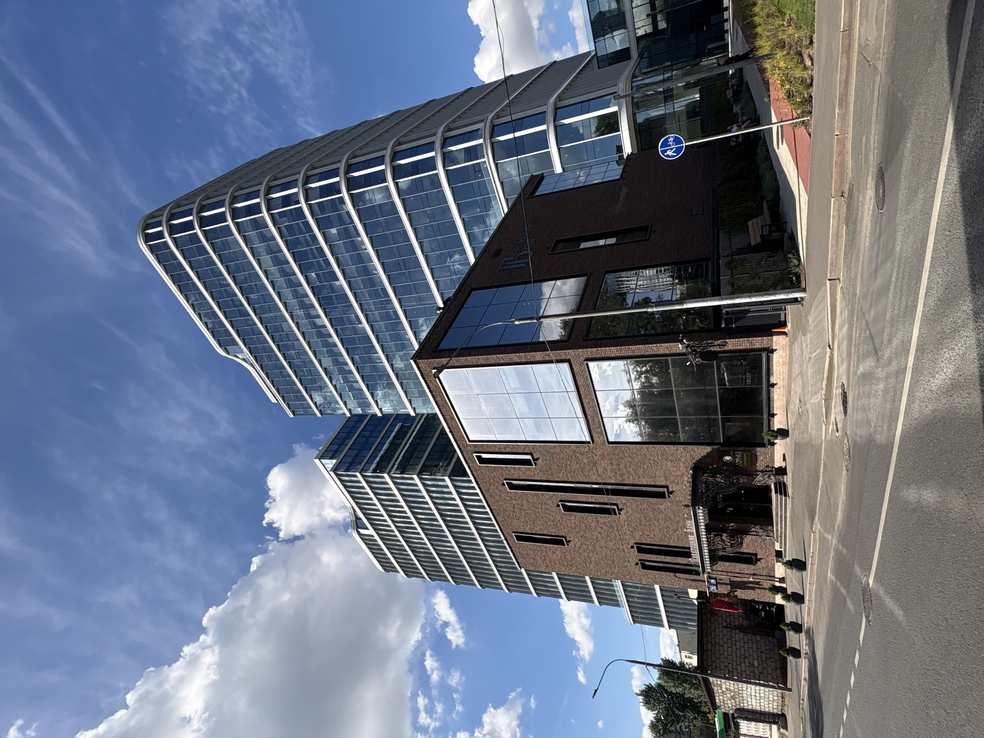 The image shows a modern architectural complex in Moscow, Russia, featuring a combination of brick and glass buildings. The foreground displays a street with asphalt paving and visible road markings. To the right, a sidewalk with a red and white tiled path runs along the buildings. A blue circular sign with icons of a pedestrian and a bicycle indicates a shared path. The main buildings consist of a lower, dark brick structure with tall, narrow windows and a more prominent, taller building with a curved facade clad in glass. The taller building's glass exterior reflects the sky and clouds. Two distinct buildings are visible behind the brick structure, one with a stepped, white-framed glass facade and another partially obscured, also with a glass exterior. The sky is a bright blue with scattered white clouds, suggesting a clear day. Trees are visible to the left of the brick building, and a street lamp stands nearby. Several small, potted trees are placed along the walkway in front of the brick building.