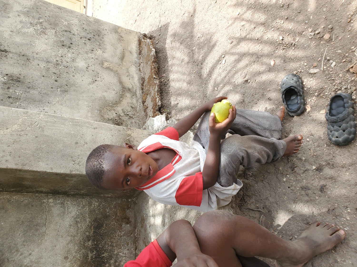 This outdoor image, taken in Mararaba, Nigeria, features a young boy sitting on the dusty ground next to concrete steps. The boy is the main subject, looking directly at the camera with a neutral and slightly curious expression. He has close-cropped dark hair and is dressed in a white t-shirt with red sleeves and collar, and grey trousers. In his left hand, he holds a light green, roundish fruit, which appears to be a guava or an unripe mango.
The setting is a sunlit, dusty area with light brown soil and scattered small stones. Shadows of leaves or branches are cast onto the ground, indicating bright sunshine, suggesting it is daytime, likely mid-morning or afternoon. To the right of the boy, a pair of dark grey, textured sandals or crocs rests on the ground. To his left, a weathered concrete structure, possibly steps or a low wall, rises.
In the lower foreground, the bare legs and feet of another person, wearing a red garment, are partially visible, suggesting companionship or an additional presence in the scene. The overall impression is of a casual, everyday moment in a modest, sun-drenched outdoor environment. There is no visible text in the image.
