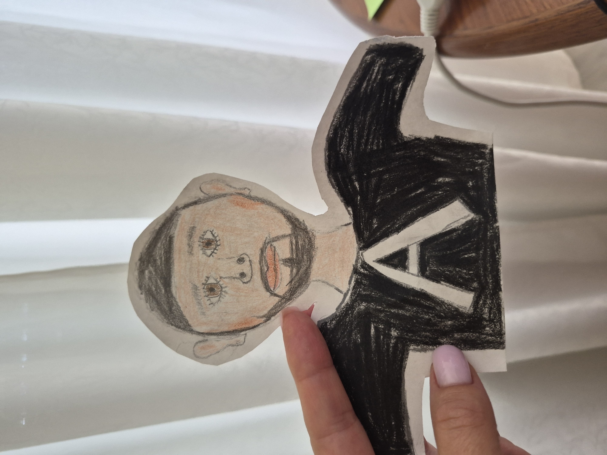 An overhead medium shot shows a cutout drawing of a person held by a hand against a white curtain background. The drawing is of a person's upper body, with a face, neck, and torso. The person is wearing a black shirt with a large white "A" on the front. The face is drawn with brown eyes, a black beard, and rosy cheeks. The background is a white curtain, which is partially lit by natural light coming from behind it. A small part of a wooden table is visible in the lower right corner.