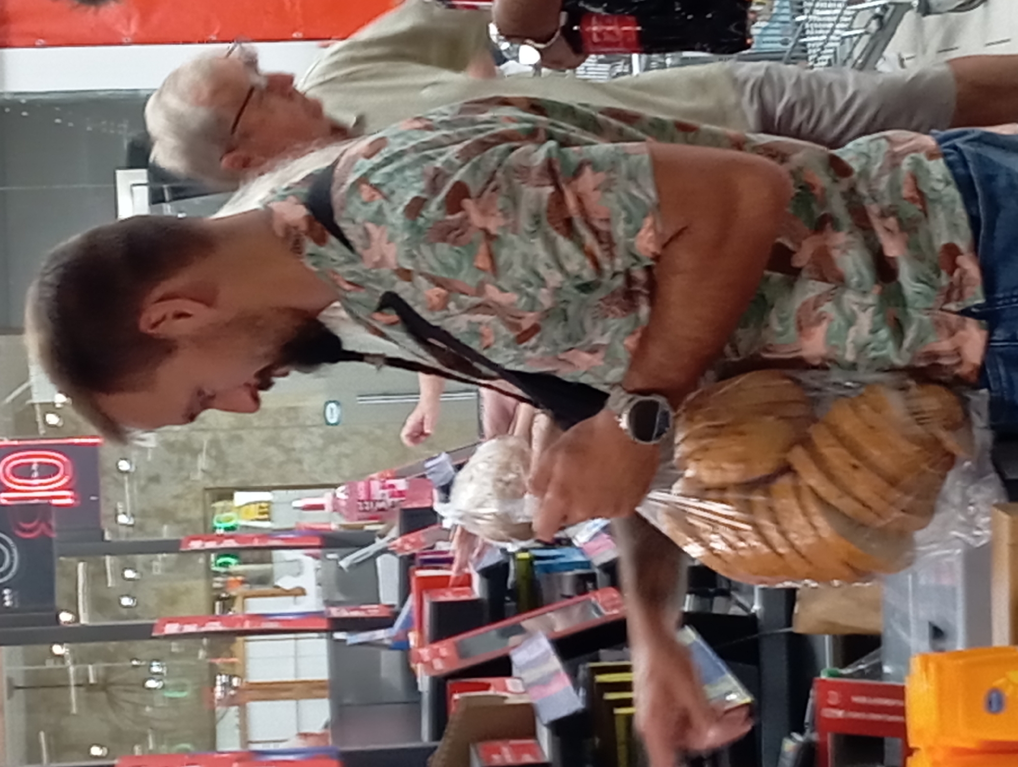 The image shows a man in a store, possibly a supermarket, in Nitra, Slovakia. He is holding a bag of sliced bread. He is wearing a short-sleeved shirt with a floral pattern and blue jeans. In the background, other people are visible, as well as shelves with products. There is a checkout counter in the foreground. A sign with the number "10" is visible on the wall in the background, suggesting this may be a checkout lane number.