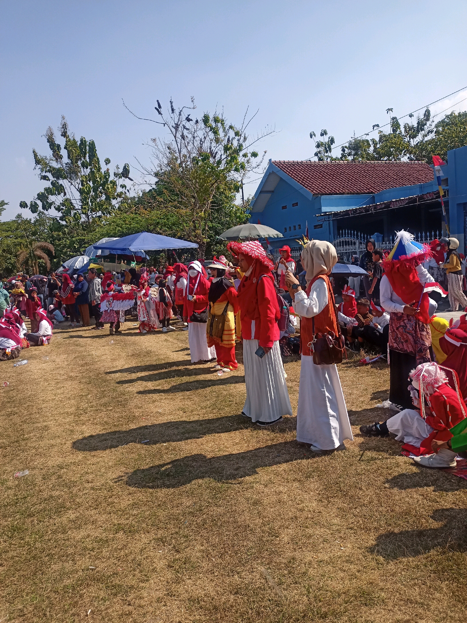 The image depicts an outdoor gathering in the city of Sumberlawang, Indonesia, likely a parade or festival, with a clear blue sky and sparse trees. A group of people, predominantly women and children, are dressed in colorful traditional or festive attire, with a prominent use of red and white, suggesting a patriotic theme. Many individuals are wearing headscarves and elaborate headdresses, and some are holding flags or small umbrellas decorated with similar colors. The ground is covered in dry, yellowed grass, with long shadows indicating the sun is casting light from the left. In the background, a blue building with a red-tiled roof is visible, along with tents and more spectators. Some people are seated on the ground, while others are standing and observing the event. The overall atmosphere appears to be one of community celebration.