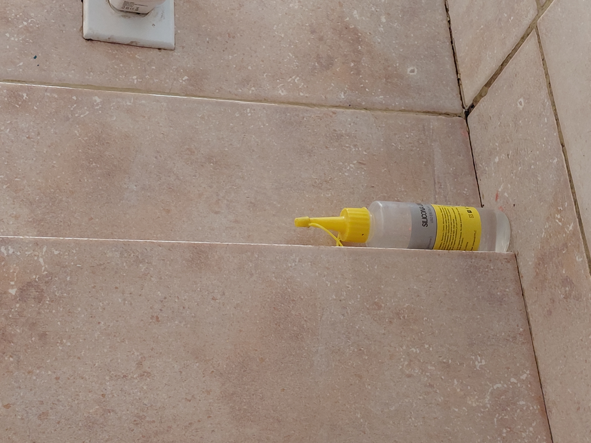 The image captures a close-up view of a tiled wall and a ledge in what appears to be an indoor setting, possibly a bathroom or kitchen in Santa Ana, Venezuela. The walls are adorned with light brown and beige speckled tiles, creating a textured, stone-like appearance. Distinct grout lines run horizontally and vertically between the tiles; some of these lines, especially in the corner, show signs of discoloration or dirt.
In the upper left quadrant of the image, a white electrical outlet is mounted on the tiled surface. Resting on a lower tiled ledge or step, positioned centrally to the right, is a clear plastic bottle. The bottle has a bright yellow, pointed nozzle and cap, and contains a translucent or white liquid or gel. A grey label on the bottle clearly displays the word "SILICONA" in black capital letters. Below this, smaller text is visible, including "USO ESCOLAR" (school use), suggesting the product's intended application. Other partial text and graphics, resembling a barcode or product codes (e.g., "ES"), are also visible on the label.
No people are present, and no active event is depicted, though the silicone bottle implies a recent or upcoming sealing, repair, or craft-related activity. The scene is well-lit, but the time of day and specific weather conditions are not discernible.