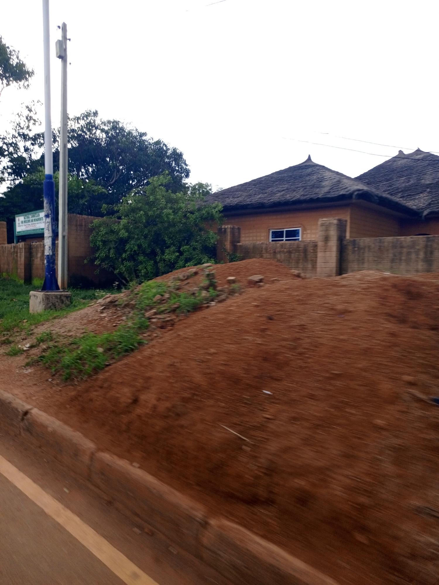 The image shows a street scene in Jos, Nigeria, with traditional architecture. In the foreground, a dirt road with a yellow lane marking runs diagonally from bottom left to top right. The ground is covered in reddish-brown soil and sparse green vegetation.
To the left, a tall, blue and white striped pole stands next to a concrete base. Behind it, a low, concrete wall encloses an area with more vegetation and some brick structures. A green sign with white text is mounted on a metal post.
Further back, a building with a thatched roof and mud-brick walls is visible. It has a small blue-paned window. Another similar building is partially visible to its right. Mature trees with dark green foliage are scattered throughout the background, creating a natural setting. The sky is overcast and bright white.